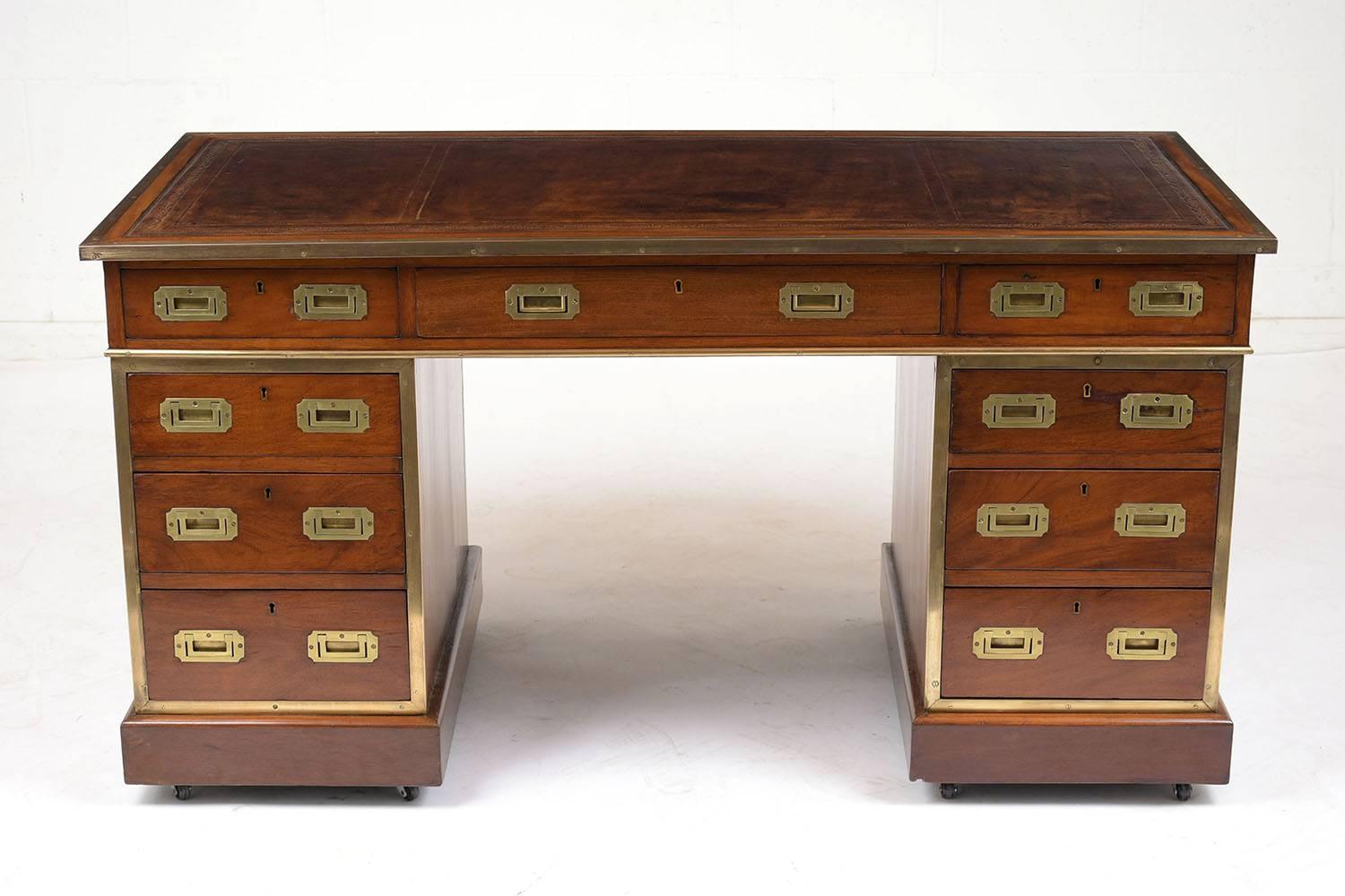 This 1900s English Campaign style pedestal desk is made of mahogany wood stained a light walnut color with a polished finish. The desk has nine drawers (four drawers on each pedestal and one pen drawer in the centre) with flush brass drawer pulls
