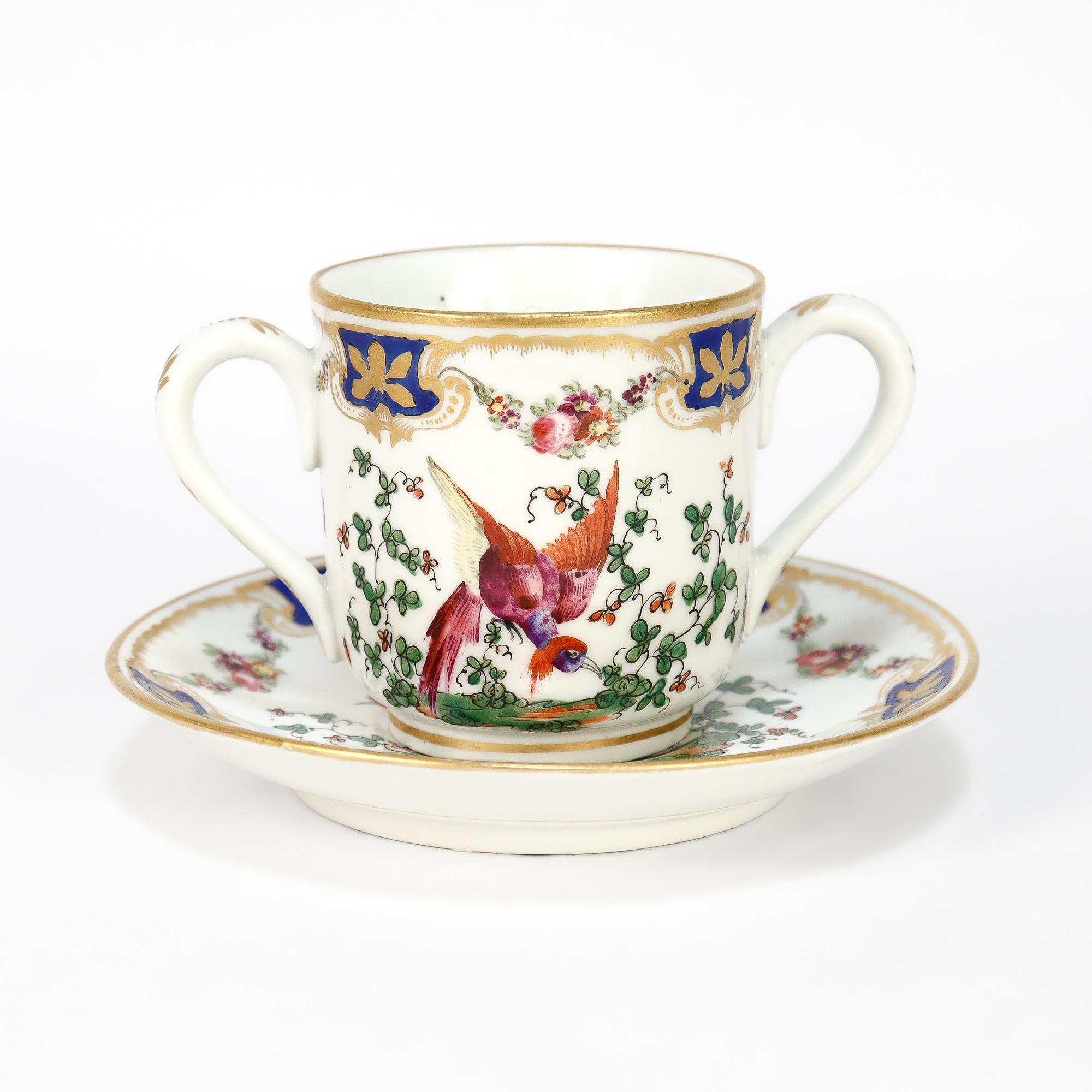 A fine antique English porcelain chocolate cup and saucer pair.

In the Chelsea Porcelain style.

Each hand painted with polychrome bird of paradise motifs surrounded by floral devices. The gilt rims accented with cobalt and gilt foliate