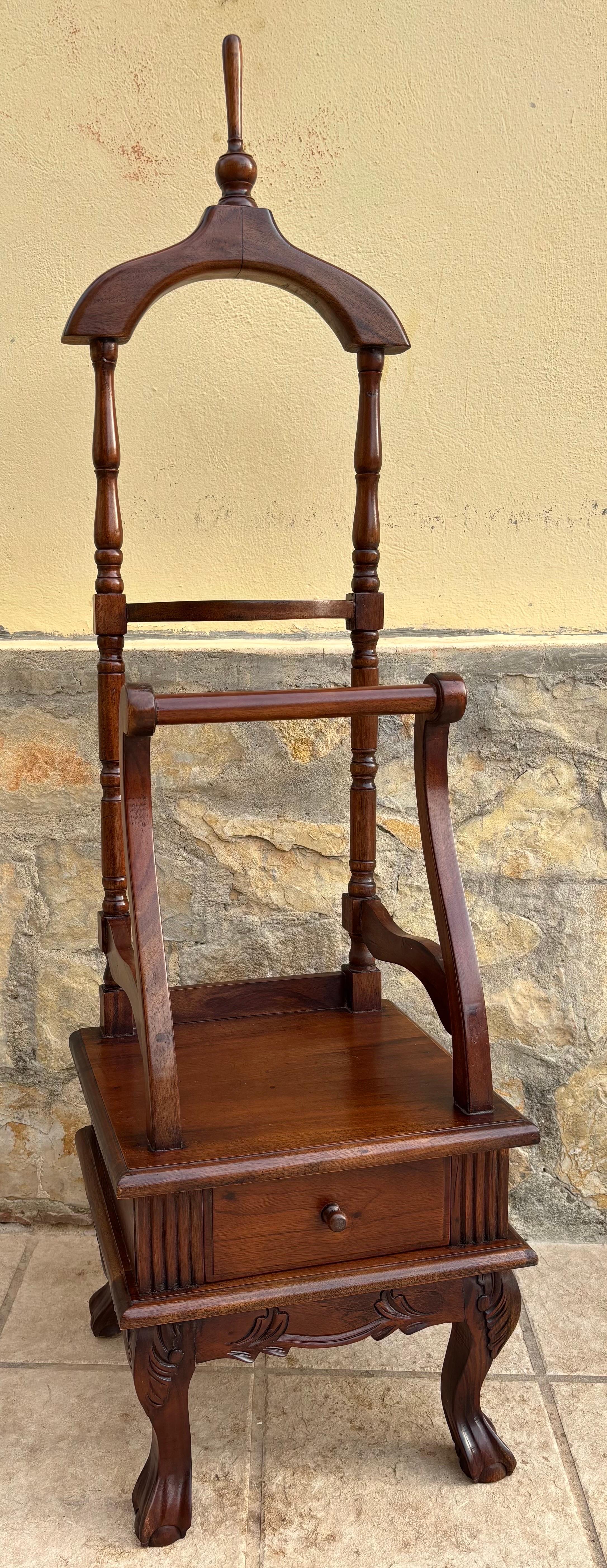 Élégant valet de chambre espagnol datant du milieu du XXe siècle, fabriqué dans un bois aux tons chauds d'acajou et à la silhouette classique et raffinée. Cette pièce allie fonctionnalité et caractère décoratif, avec un dessus de cintre sculpté, un