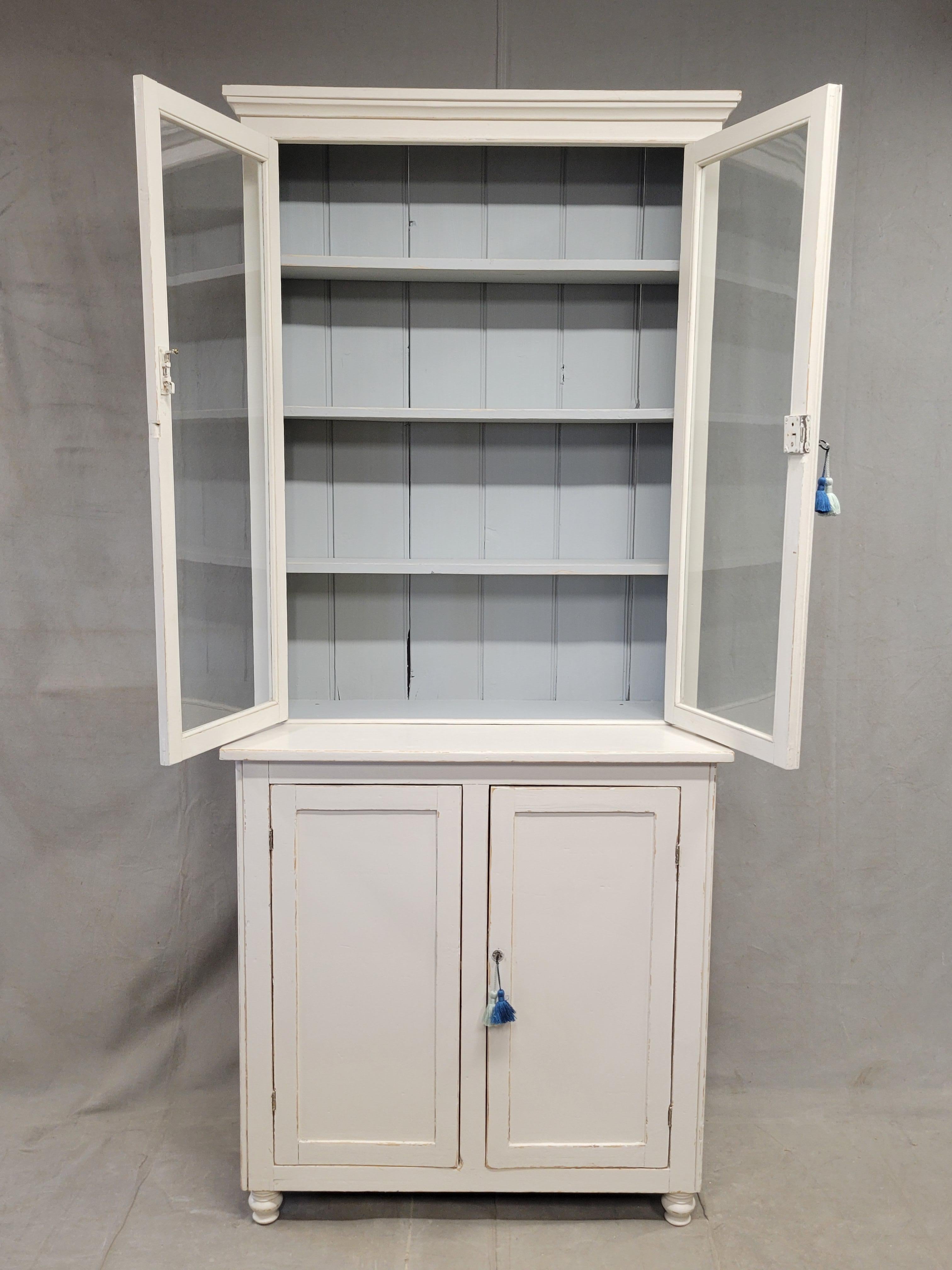 Un bellissimo e classico mobile antico degli anni '20 dipinto a mano in English Country Antiques con ante in vetro sulla parte superiore e ante massicce sulla parte inferiore. La credenza è dipinta di bianco crema... una vecchia vernice bianca che è