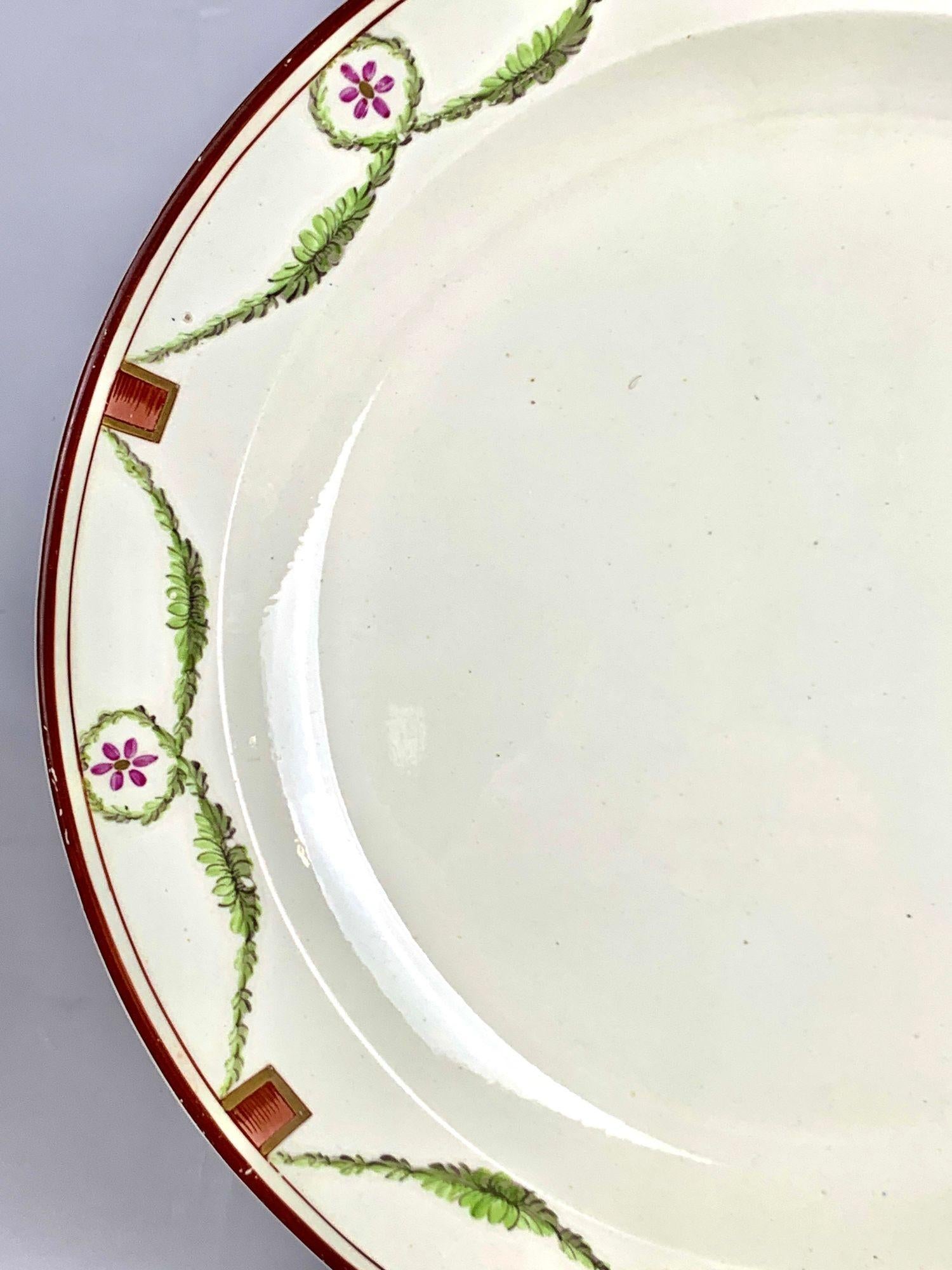 Questo bellissimo piatto antico inglese in crema, risalente al 1785, presenta un disegno di ghirlanda neoclassica dipinto a mano intorno al bordo.
Delicati fiocchi verdi circondano piccoli fiori viola. Ogni borsa è legata con dispositivi a nastro
