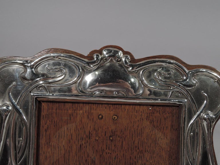 Edwardian Art Nouveau sterling silver picture frame. Made by William Aitken in Birmingham in 1905. Rectangular window in shaped scrolled surround mounted to stained wood with hinged support. Chased Japonesque ornament, cranes, cattails, and lily