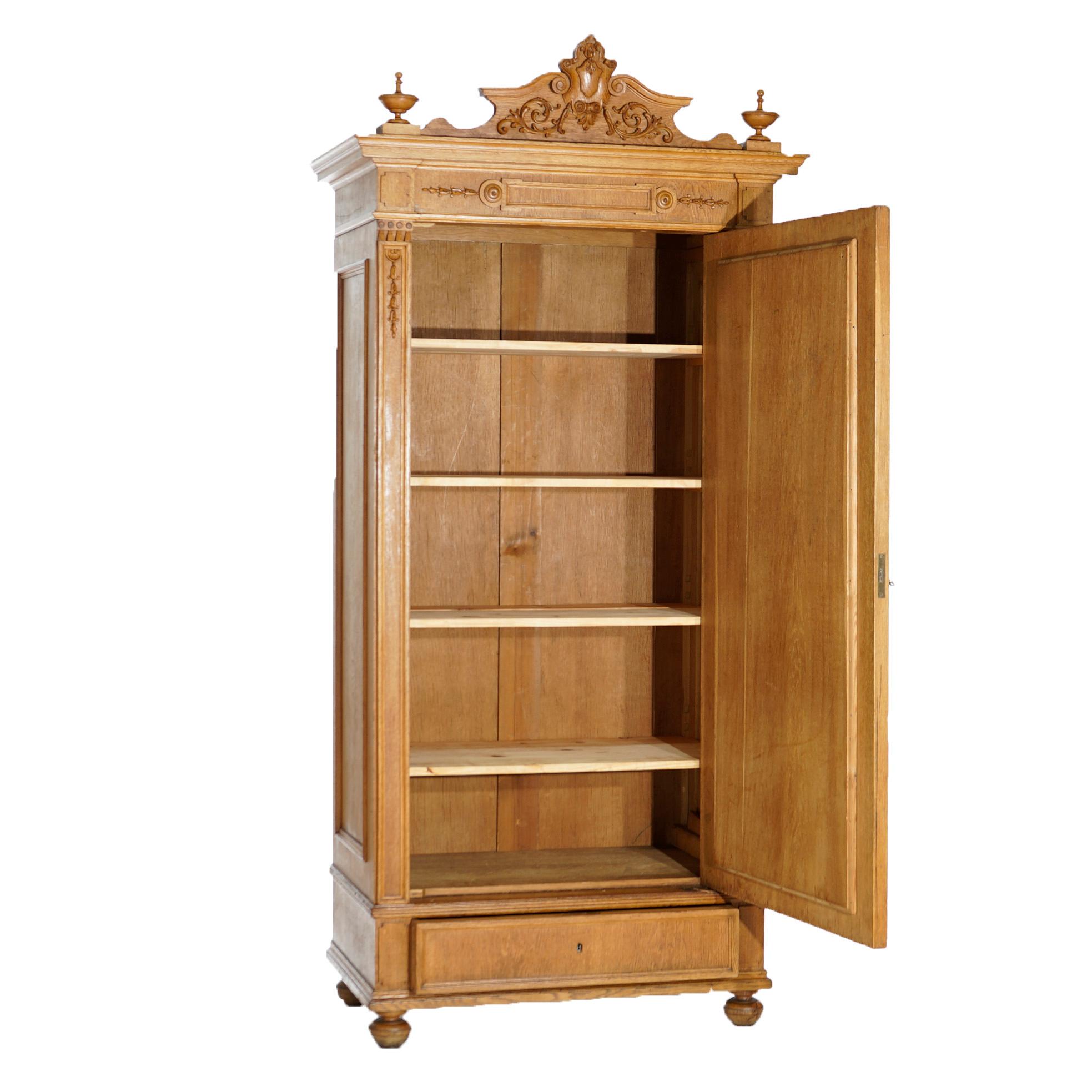 An antique English Edwardian armoire offers oak construction with carved crest having central shield and flanking foliate and scroll elements over case with single mirrored door opening to shelved interior over single long drawer, raised on