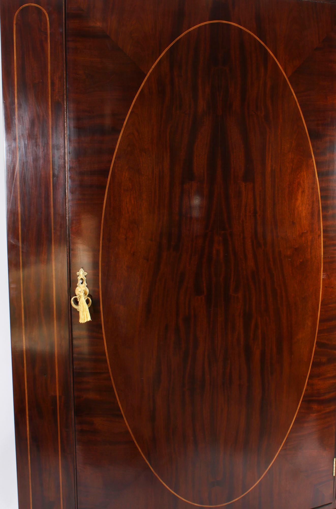 This is an impressive English antique Edwardian Sheraton Revival bowfront flame mahogany wardrobe, circa 1900 in date.

It has a decorative flared key carved cornice with a crossbanded frieze over a single oval satinwood inlaid door enclosing a