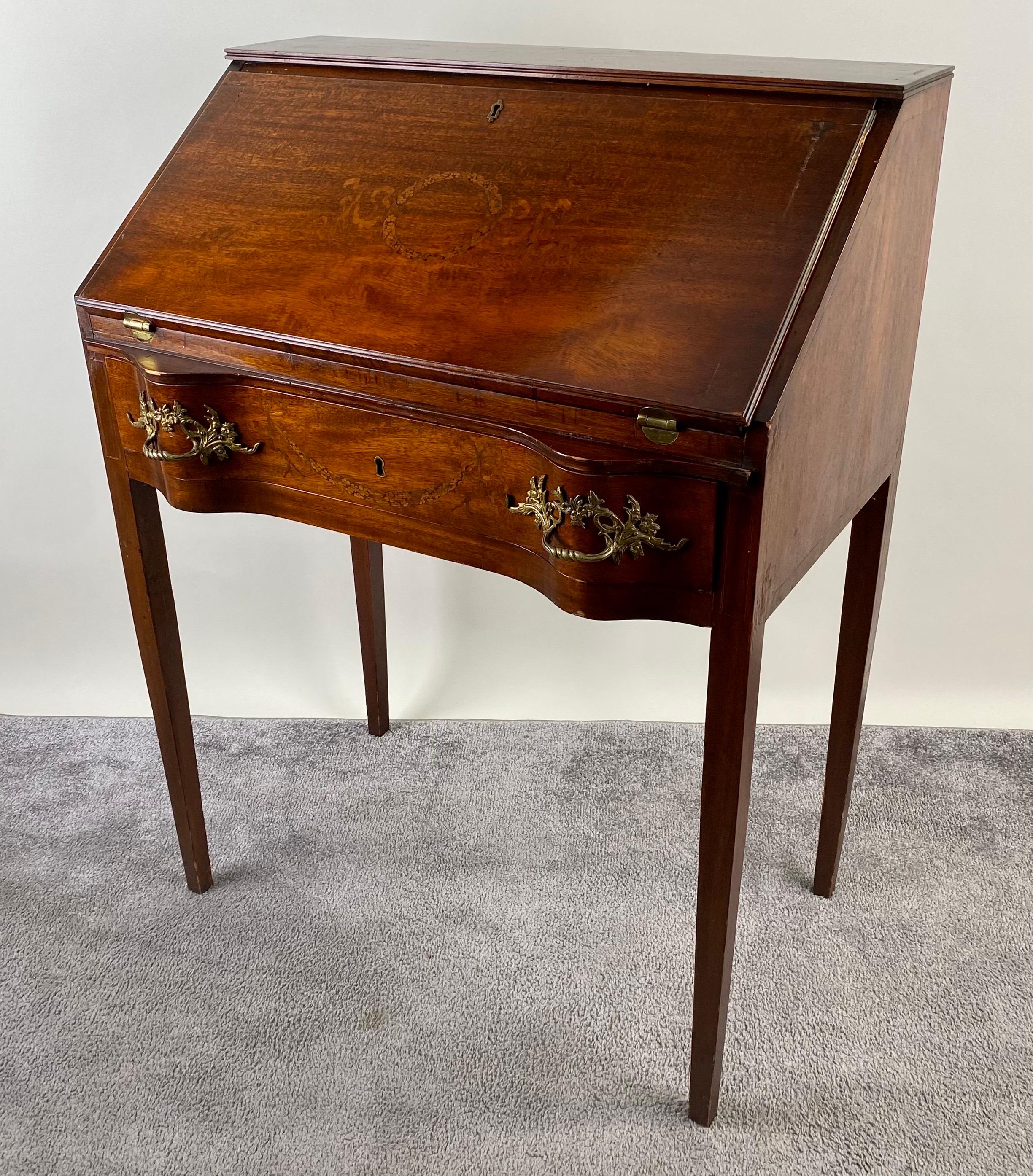 An antique English Edwardian slant front writing desk, a true embodiment of the refined craftsmanship of the early 1900s. Crafted from rich mahogany, the focal point of this masterpiece lies in the intricate marquetry inlay gracing the drop-front of