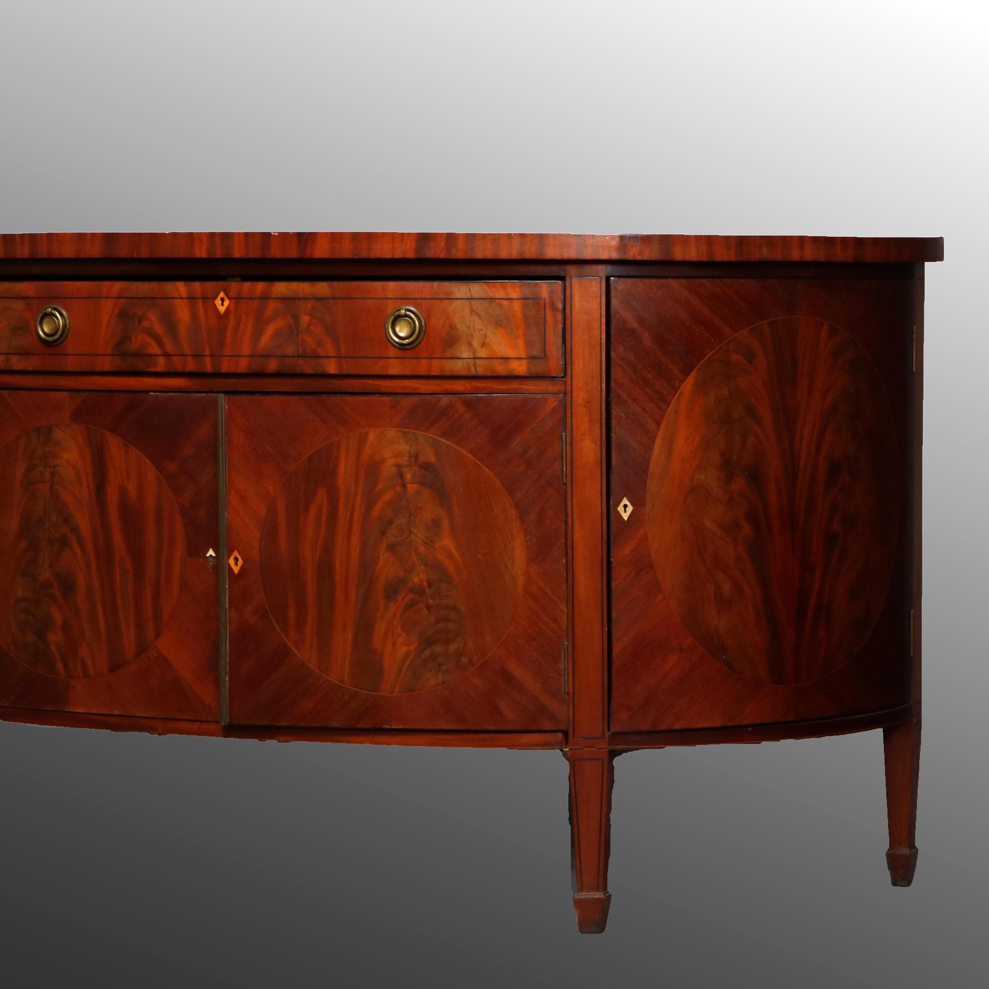 An English George III sideboard offers deeply striated mahogany construction in demilune form with bookmatched frieze drawer surmounting double door cabinet and flanked by quarter round shelved cabinets, ebonized banding, inlaid diamond escutcheons