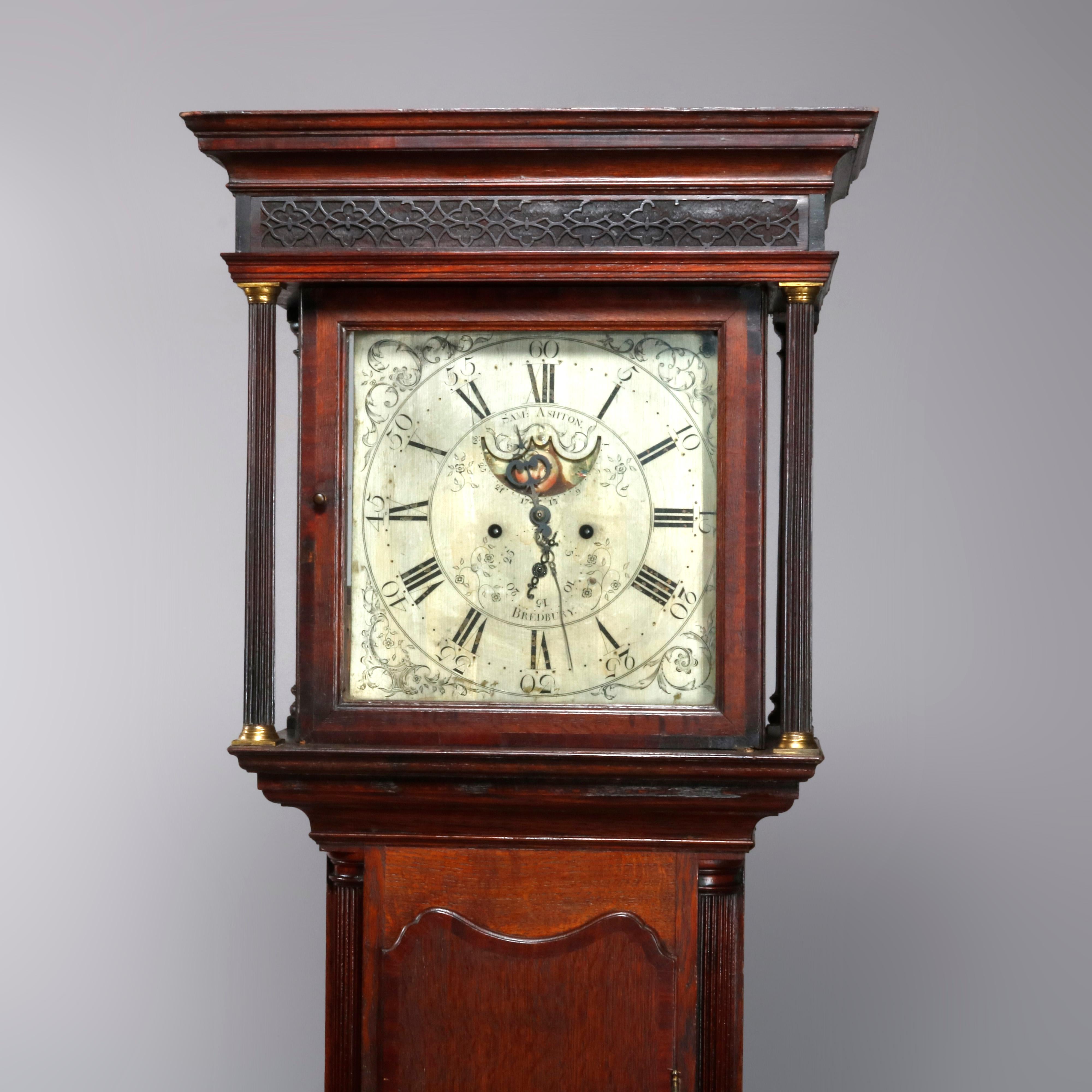 An antique George III longcase clock offers English oak construction with moulded cornice hood with reeded pilasters having bronze capitals and bases; porcelain foon phase face with Roman numerals and signed Sam Ashton, Bredbury, 18th