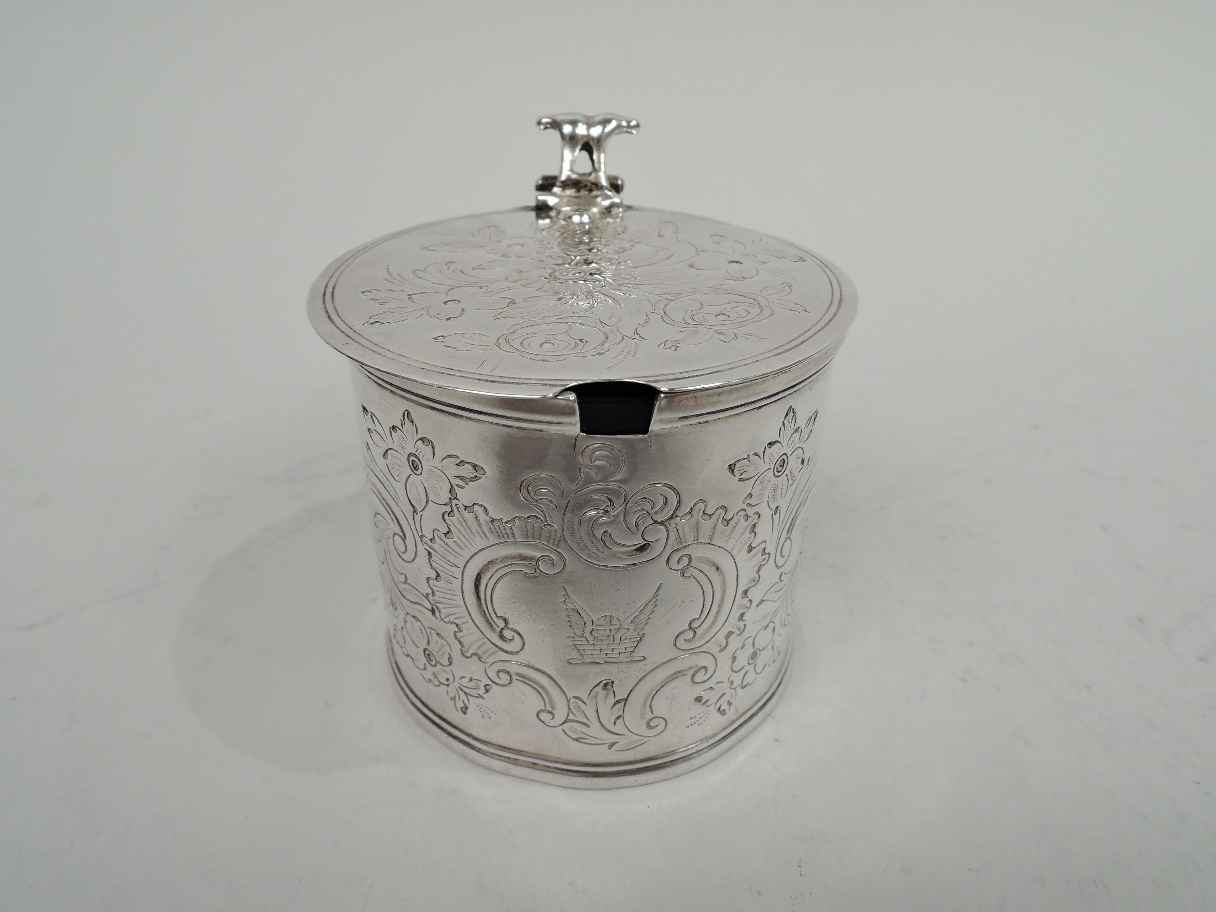 George III sterling silver mustard pot. Made by Andrew Fogelberg in London 1770. Drum-form with flat and hinged cover; leaf-capped s-scroll handle with open scroll thumb rest. Engraved leafing scrollwork, including frame engraved with armorial.