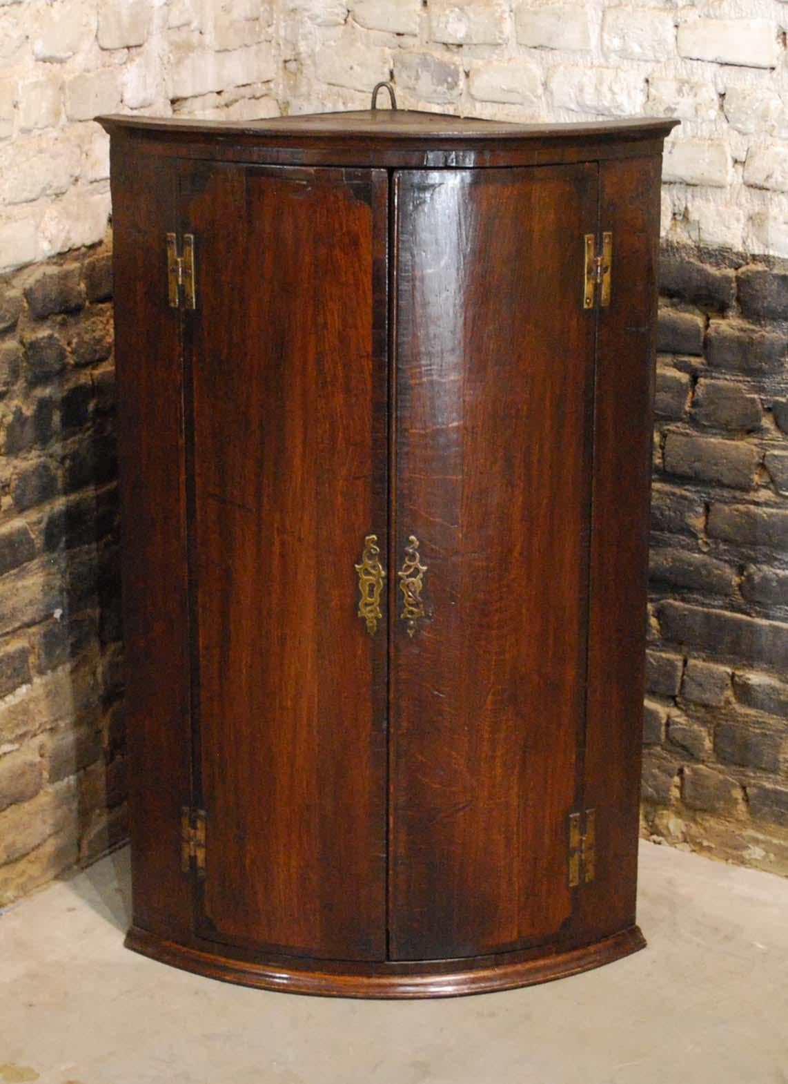 This elegant antique hanging corner cabinet was made in England circa 1860. 
It is made in solid oak with crossband mahogany veneer surrounding both bow fronted doors. It’s interior features three fixed shelves and has a warm dark brown painted