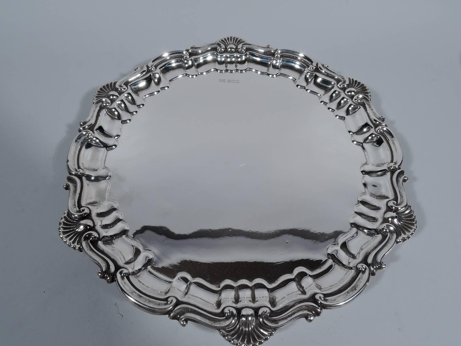 George V sterling silver salver. Made by Walker & Hall in Sheffield in 1916. Circular with tapering sides and molded rim: alternating s- and c-scrolls interspersed with scallop shells. Rests on three scrolled supports. Hallmarked. Weight: 17.5 troy
