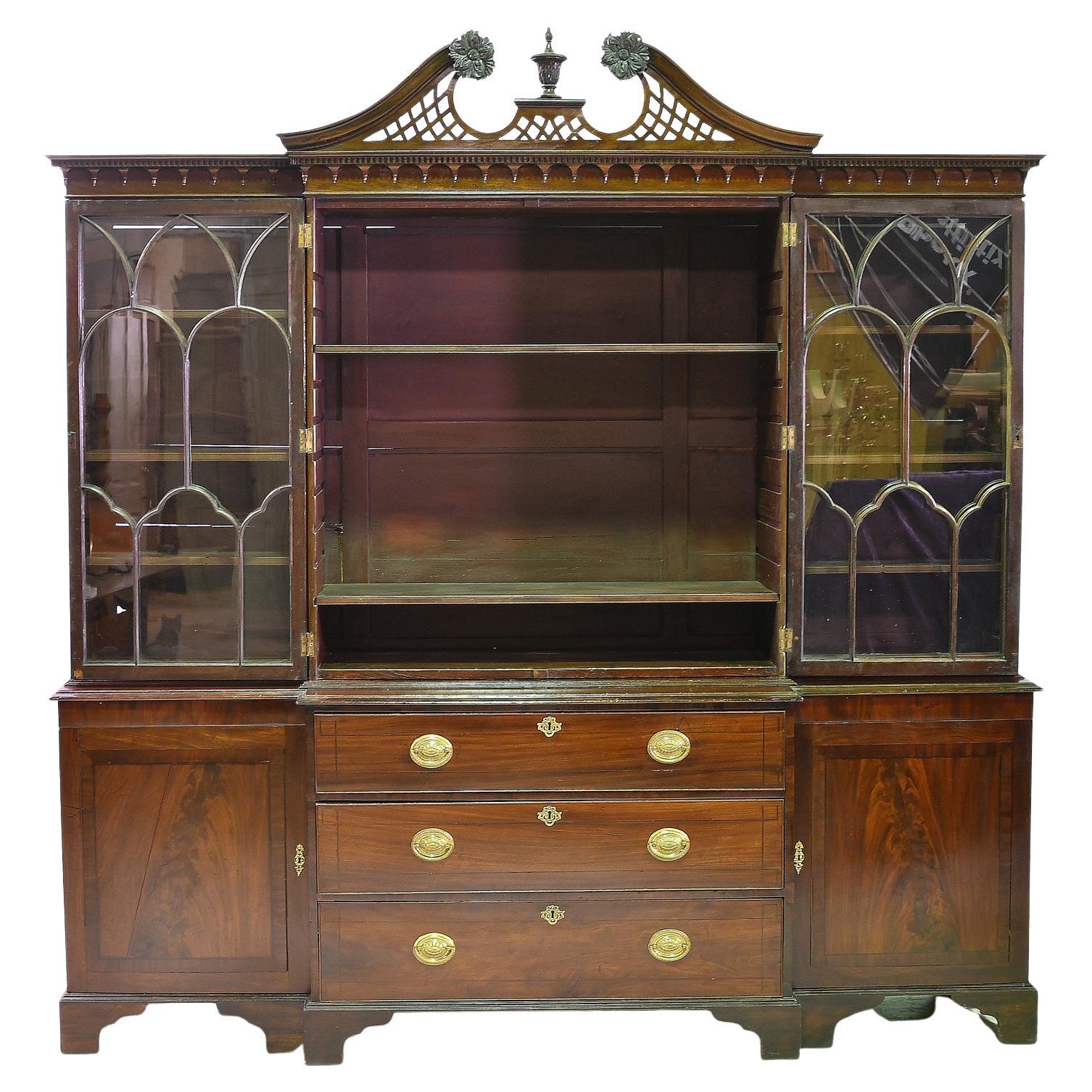 A handsome Georgian-style breakfront bookcase in fine mahogany with arched mullioned glazed panels & a scrolled lattice pediment top with dentil crown molding, below which are carved pendants. The projecting center section offers three storage