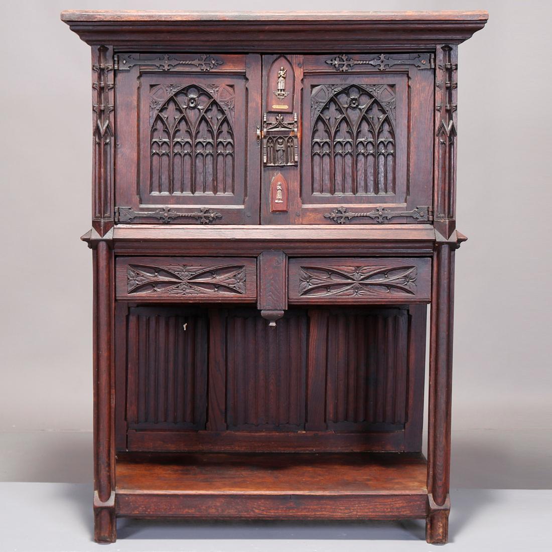 An antique English Gothic court cupboard offers oak construction with heavily carved double doors having decorated arch reserves and opening to shelved interior surmounting two lower drawers, bronze hardware throughout, 19th century

Measures: 52