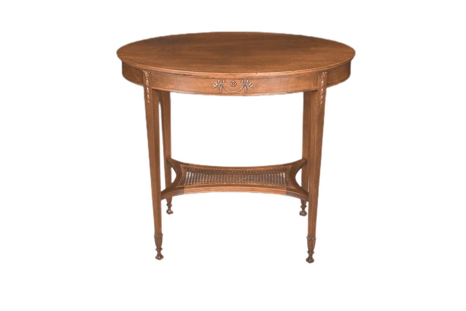 Early 20th century George Hepplewhite style oval carved mahogany two tiered accent or occasional table with caned shelf, circa 1930s. It is finished and carved on all sides so it can be placed in the center of a room or against a wall. It has a