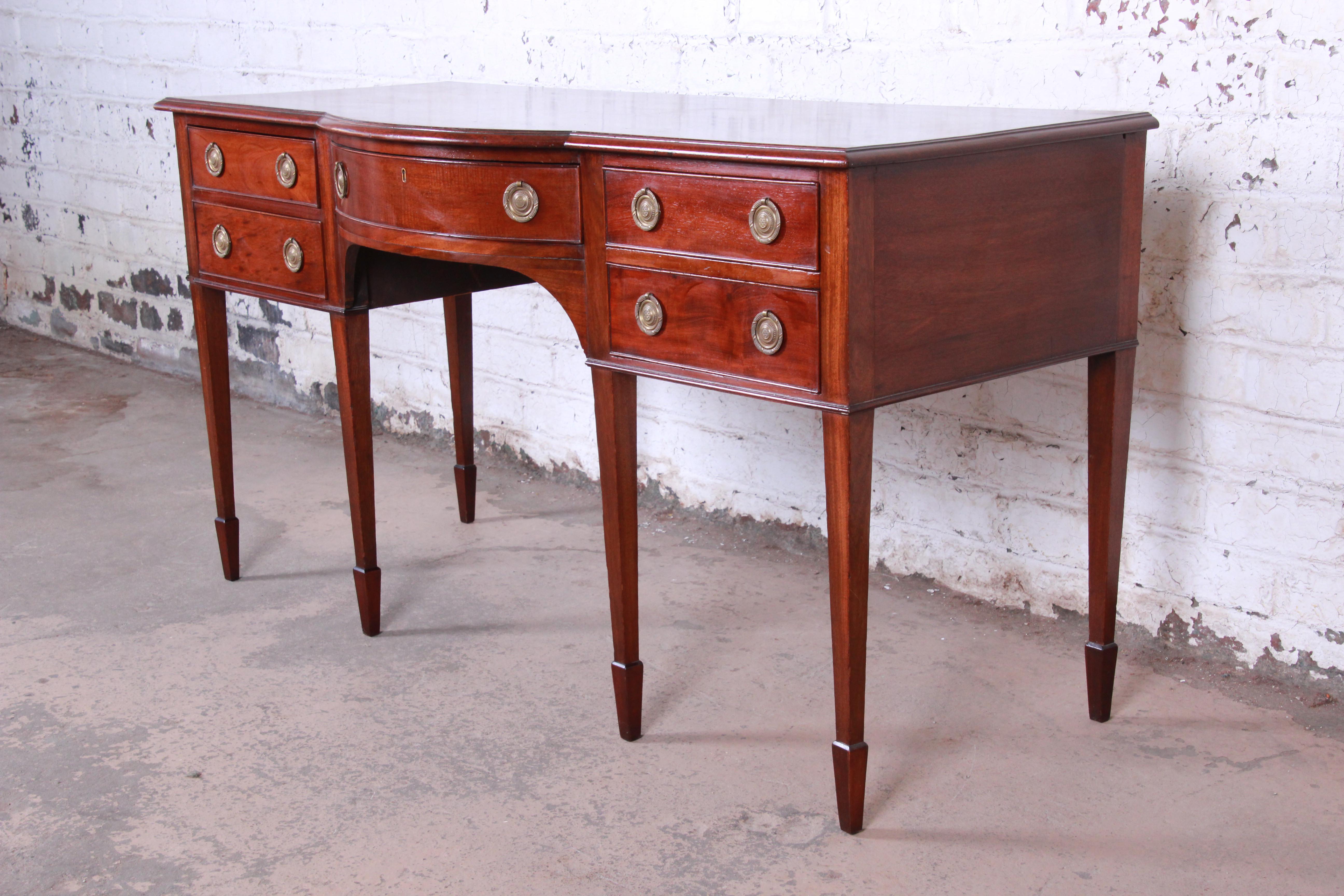 A gorgeous antique Hepplewhite style mahogany sideboard or buffet server. The sideboard features beautiful mahogany wood grain and original brass hardware. It offers good storage, with five dovetailed drawers. The case rests on tall legs with spade