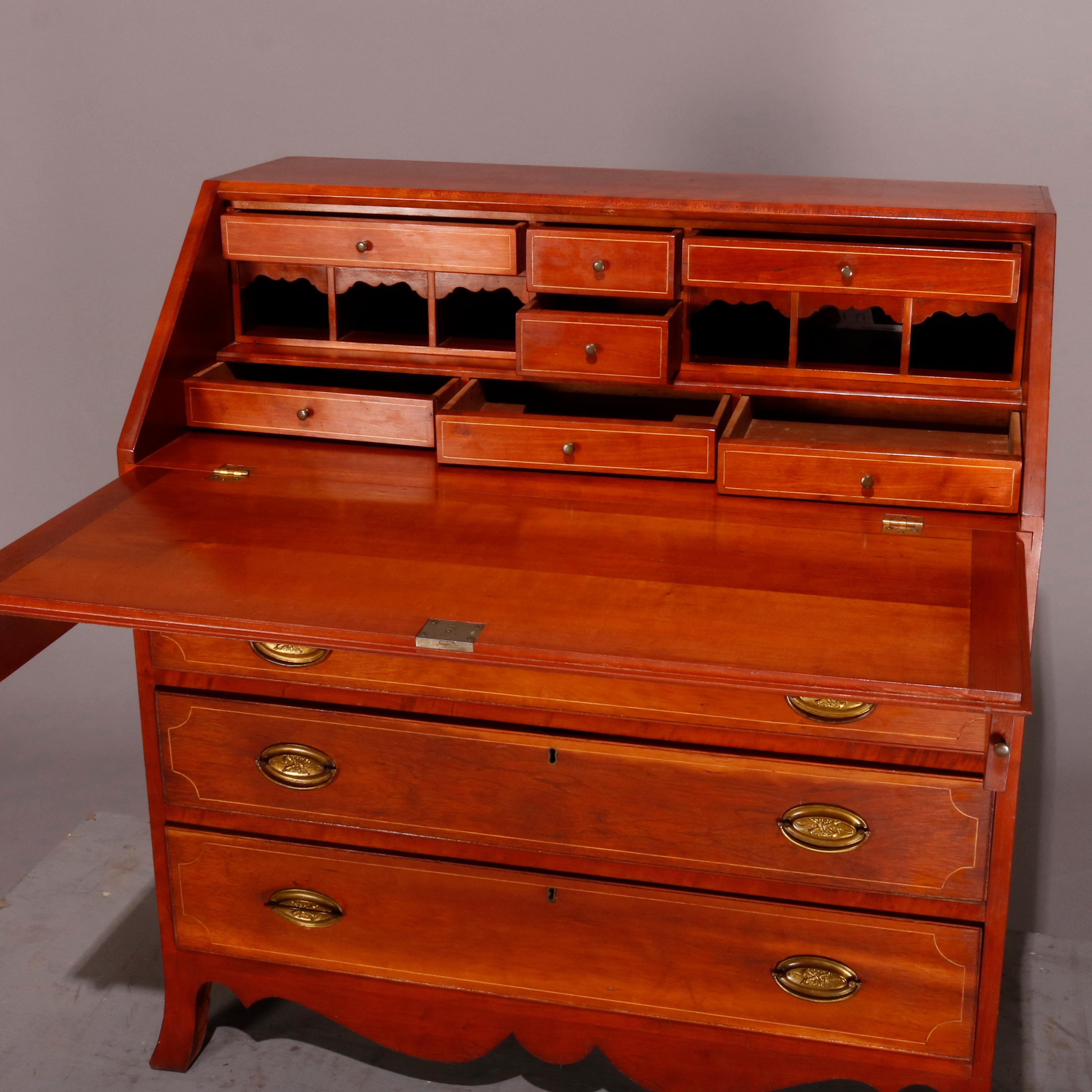 An antique Hepplewhite style slant front desk offers cherry construction with drop front opening to reveal writing surface surmounted by smaller drawers and pigeon holes and supported by pull-outs, case with four long drawers each having cast bronze
