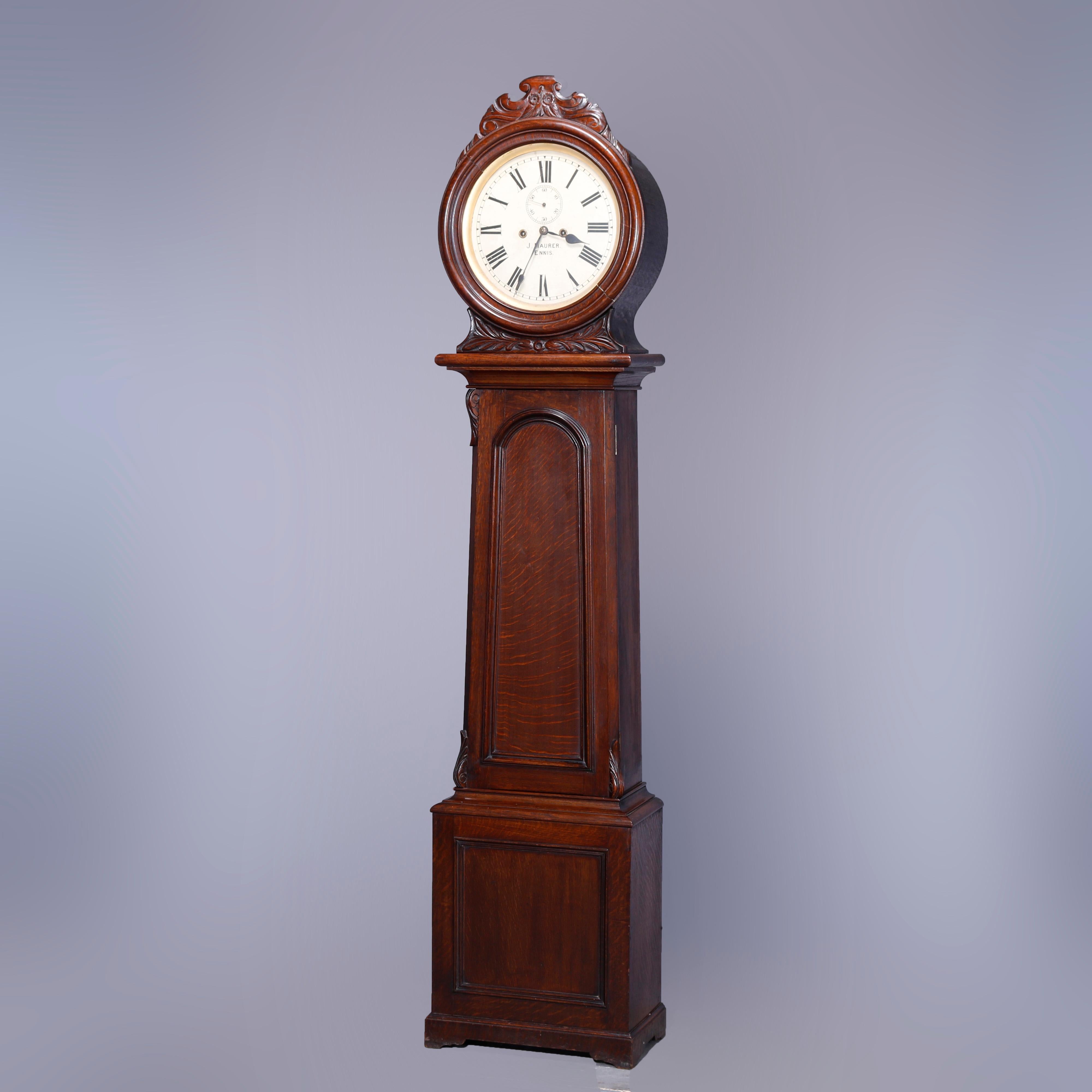 An antique English tall case clock by J. Maurer, Ennis offers oak case with circular hood having carved foliate elements surmounting flared and paneled trunk raised on bracket feet, maker or retailer signed on face, weight driven, complete and not