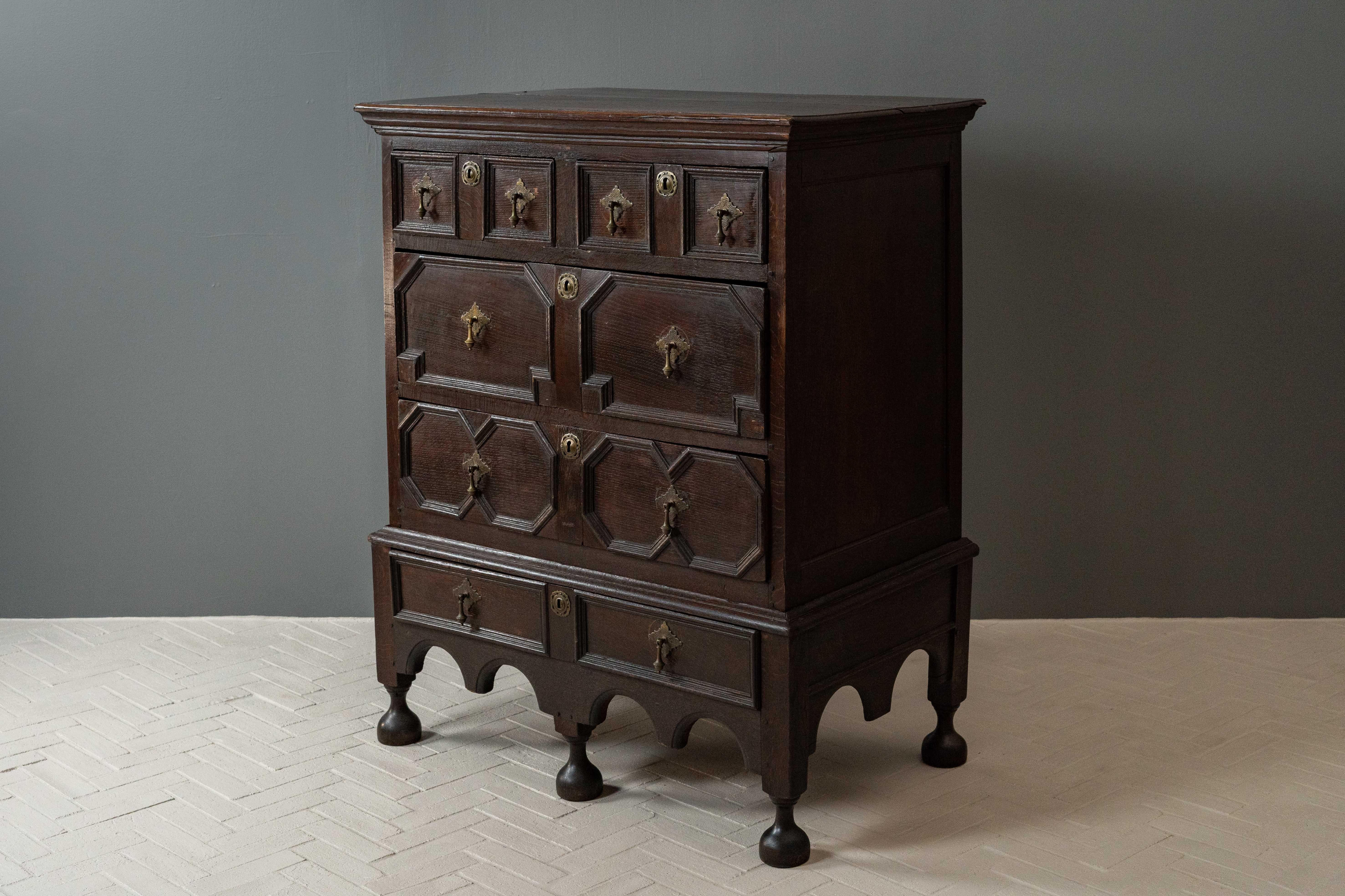 England, ca. late 17th century. A fabulous antique Jacobean or Charles II style chest on stand. Features solid oak construction with a planked top with moulded edge above two small and two large geometrically paneled drawers. Cast brass drop handles