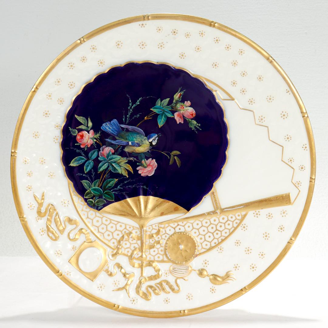 A fine antique English Aesthetic Movement porcelain plate.

Attributed to Bodley.

With a trompe l'oeil decorated Japanese fans (an uchiwa and a sensu) & precious objects to the plate. The uchiwa fan is painted and enameled with roses & a blue