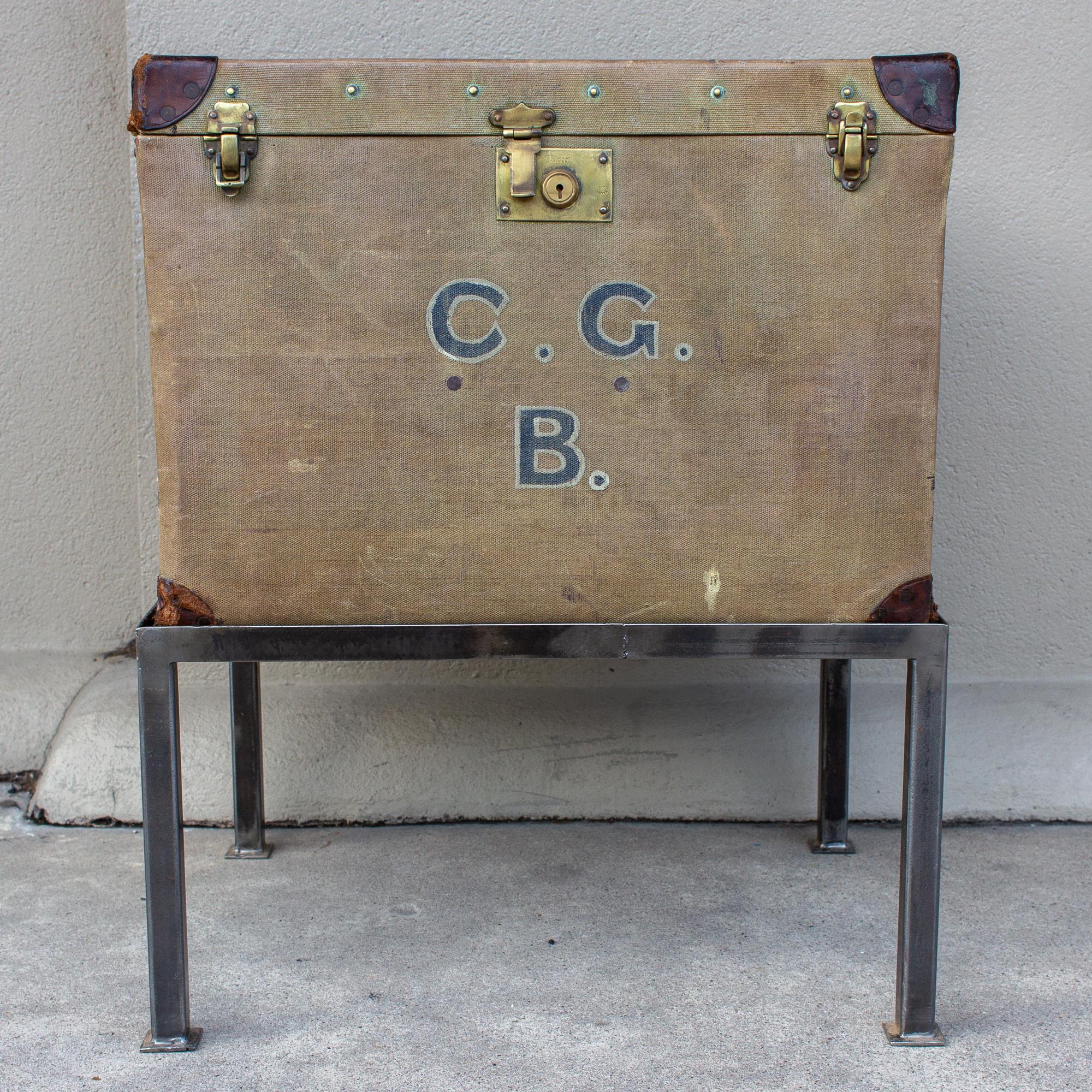 This antique English luggage piece has been crafted into a side table with a custom iron base. The trunk has the initials CGB on the front and was part of a luggage set belonging to an officer in the British Royal Navy. The trunk is a khaki green