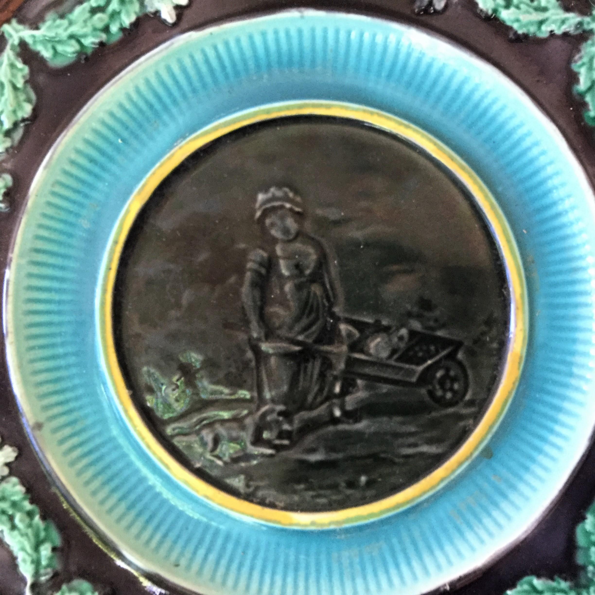 19th century English Majolica plate featuring a woman with a dog pushing a wheelbarrow bordered with a garland of oak leaves. No maker's mark.