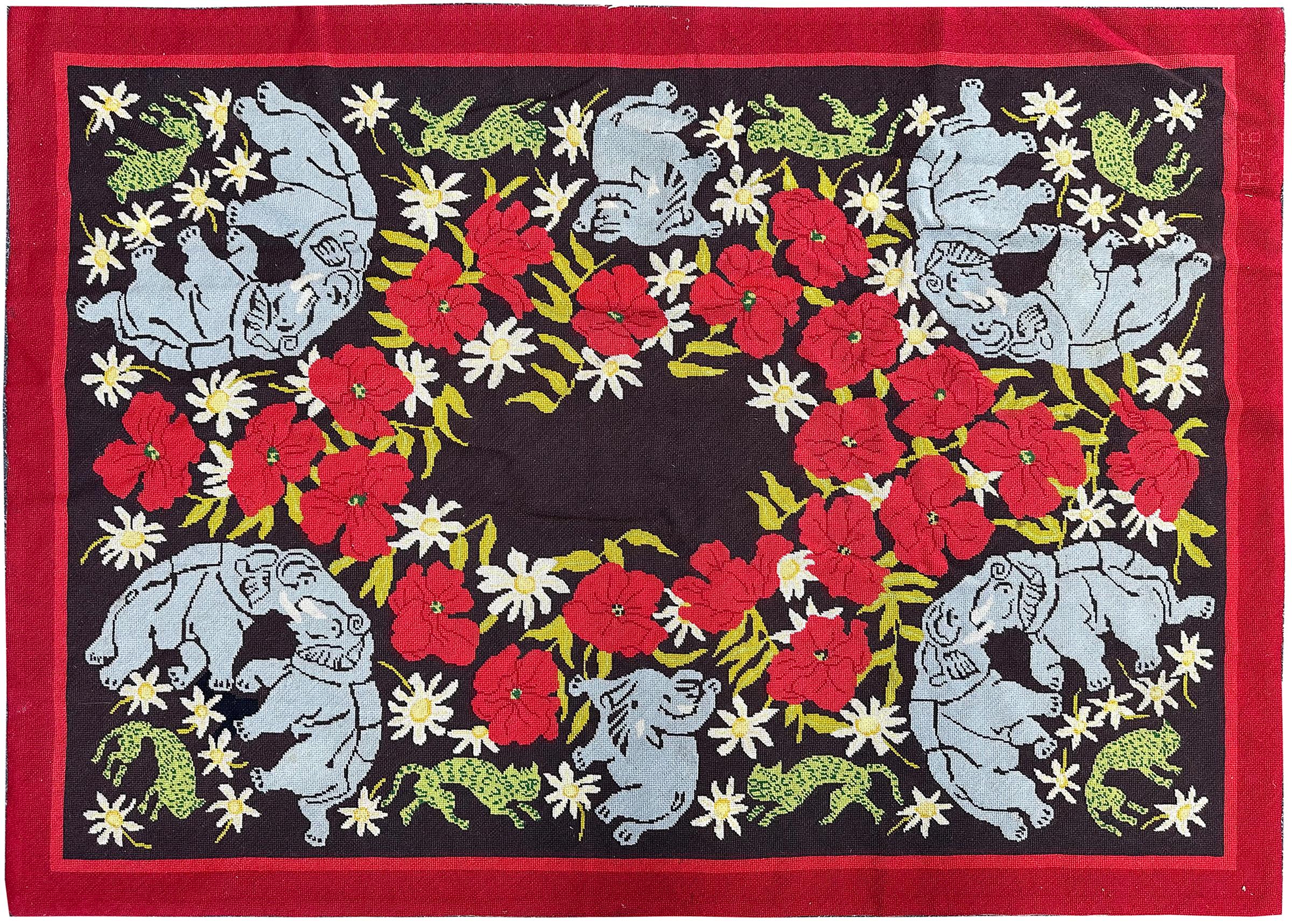 This charming English needlepoint tapestry from Antique Rug Collection features a whimsical ring of blue elephants surrounded by bold red florals, white daisies, and playful green animals set against a dark ground. The lively color palette and
