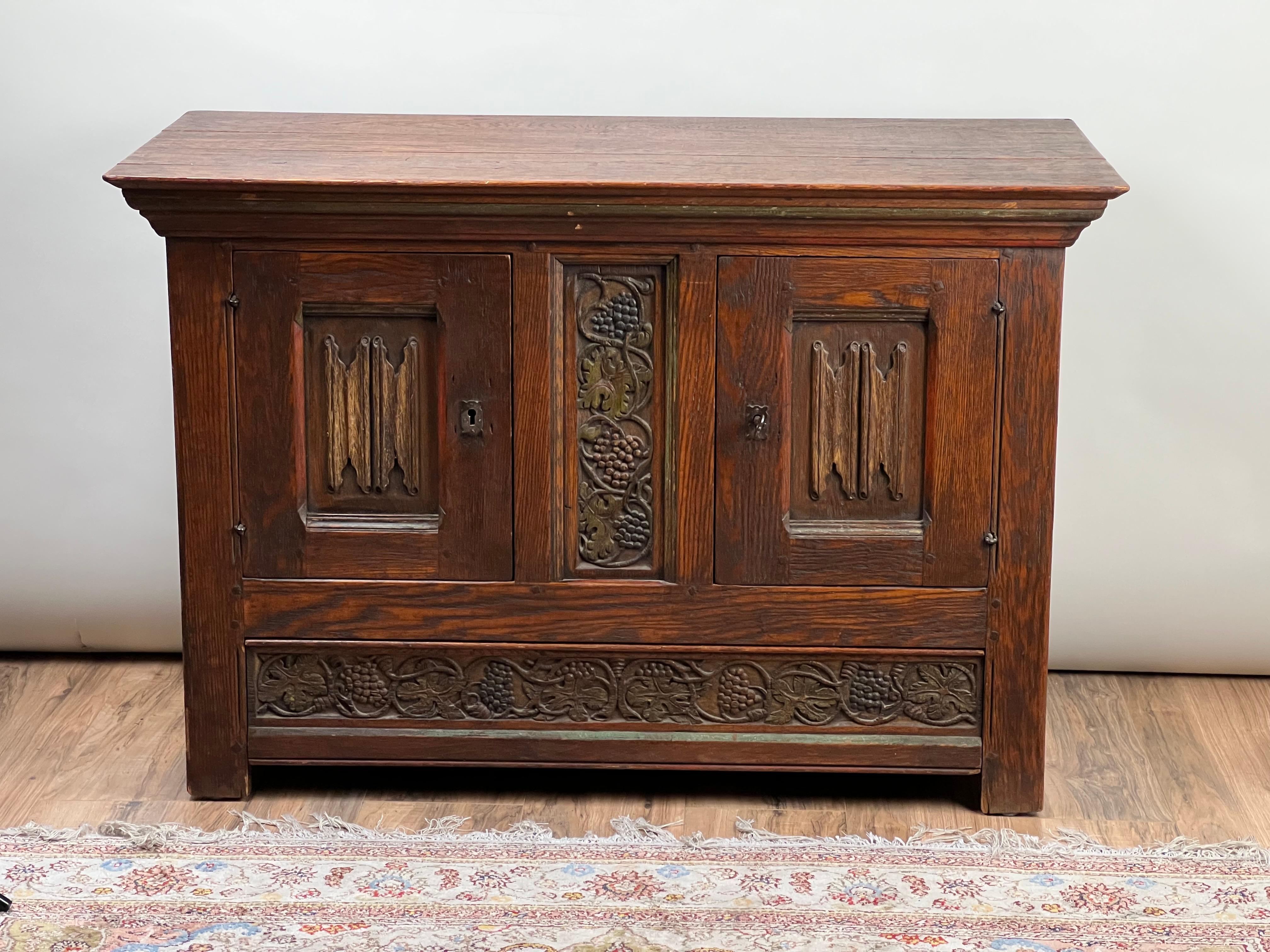 This Antique English Oak Court Cabinet exudes history and craftsmanship, featuring intricately carved linen fold panels and bold detailing. The center door panel is adorned with a grapevine motif, symbolizing abundance and prosperity, beautifully