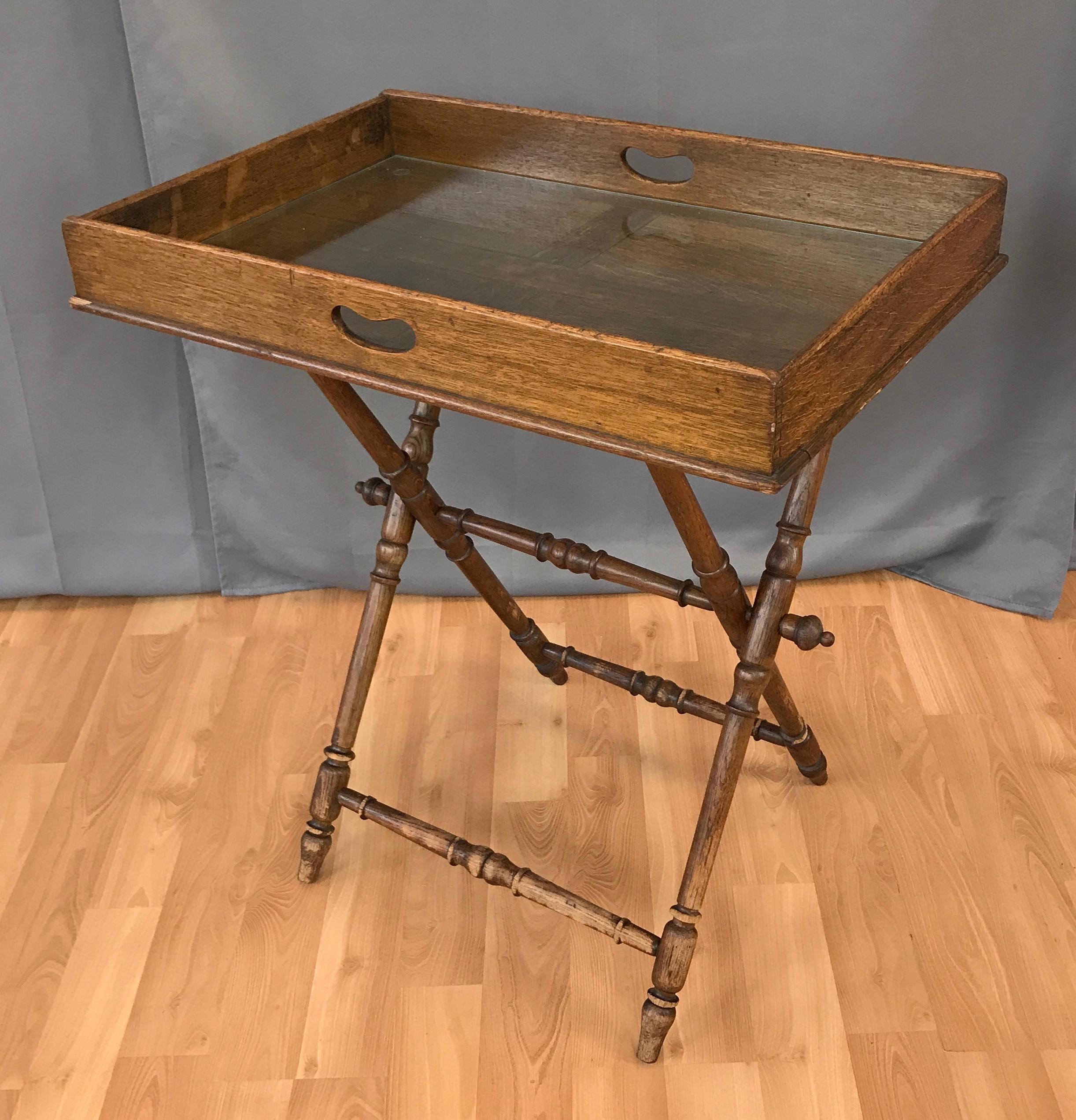 Antique English oak butlers tray on folding base from the Victorian period. 

The removable tray features keyhole side handles, exposed dovetail joinery and a glass insert to protect the tray surface.
The Campaign style folding base has