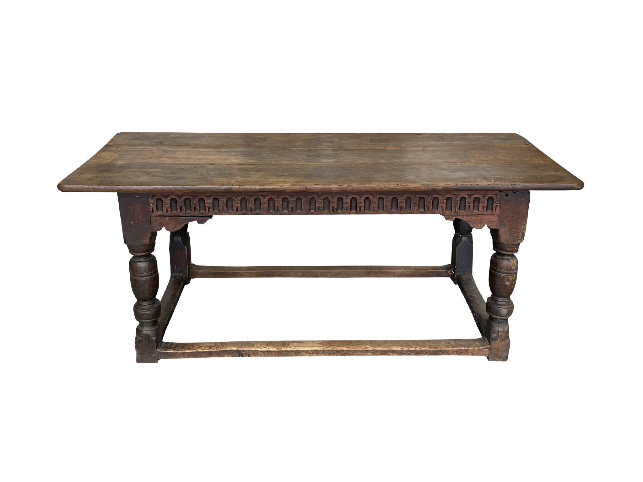 Crafted in the Late 18th-Early 19th Century, this English table is comprised of oak. Hand-carved designs adorn the skirt. The legs have a turned and block form design and are held by stretchers. The top was a later replacement, but is still antique.