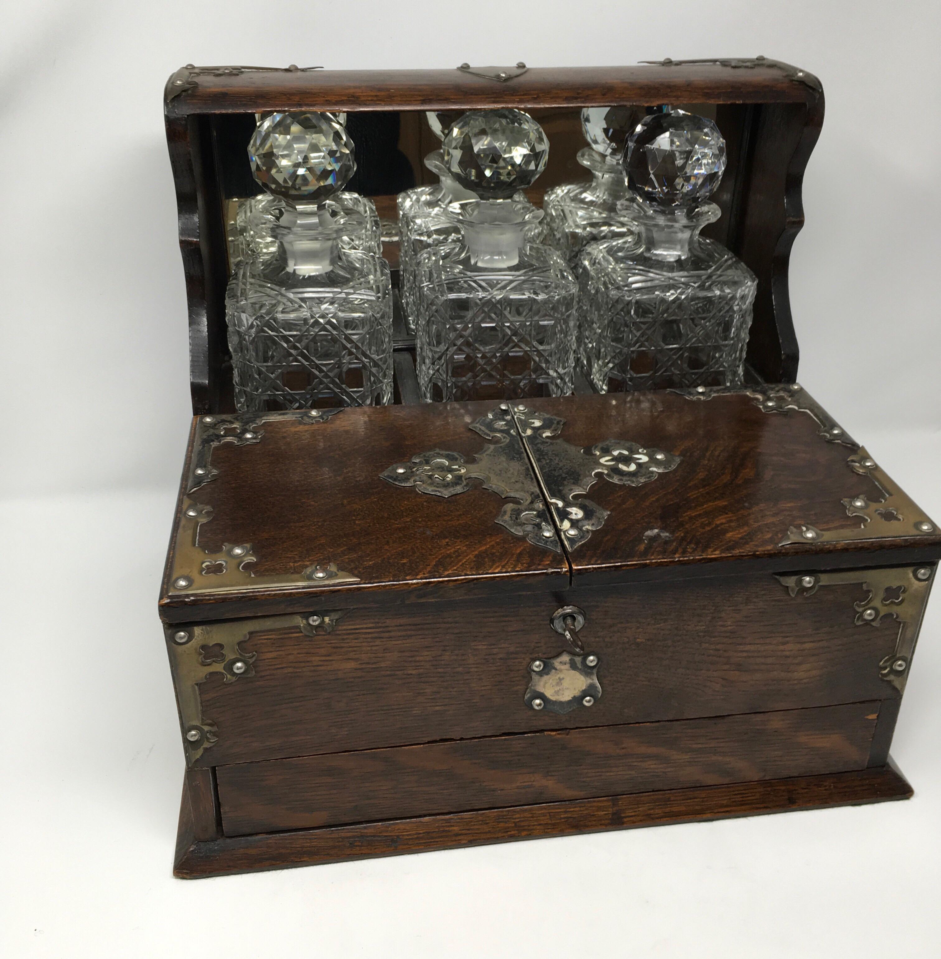 This is an antique English oak Tantalus with three decanters and a hidden games drawer. The three square decanters are in good condition although one has a small chip on the inside of the stopper. The back of the decanter section is mirrored and the