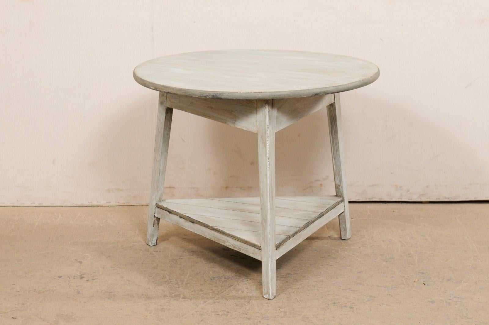 An English painted wood center table with shelf from the early 20th century. This super cute antique table from England has a round-shaped top, which rests above a triangular-shaped apron, with a triangular stretchered lower shelf beneath, which is