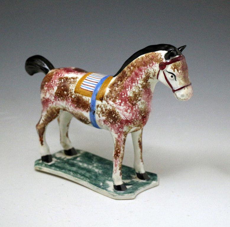 Pottery pearlware figure of a saddled horse standing on a thin green mottled coloured base. 
The horse is sponge decorated in shades of maroon and brown a pleasing effect with the mustard , maroon and blue striped saddle with its blue strap. The