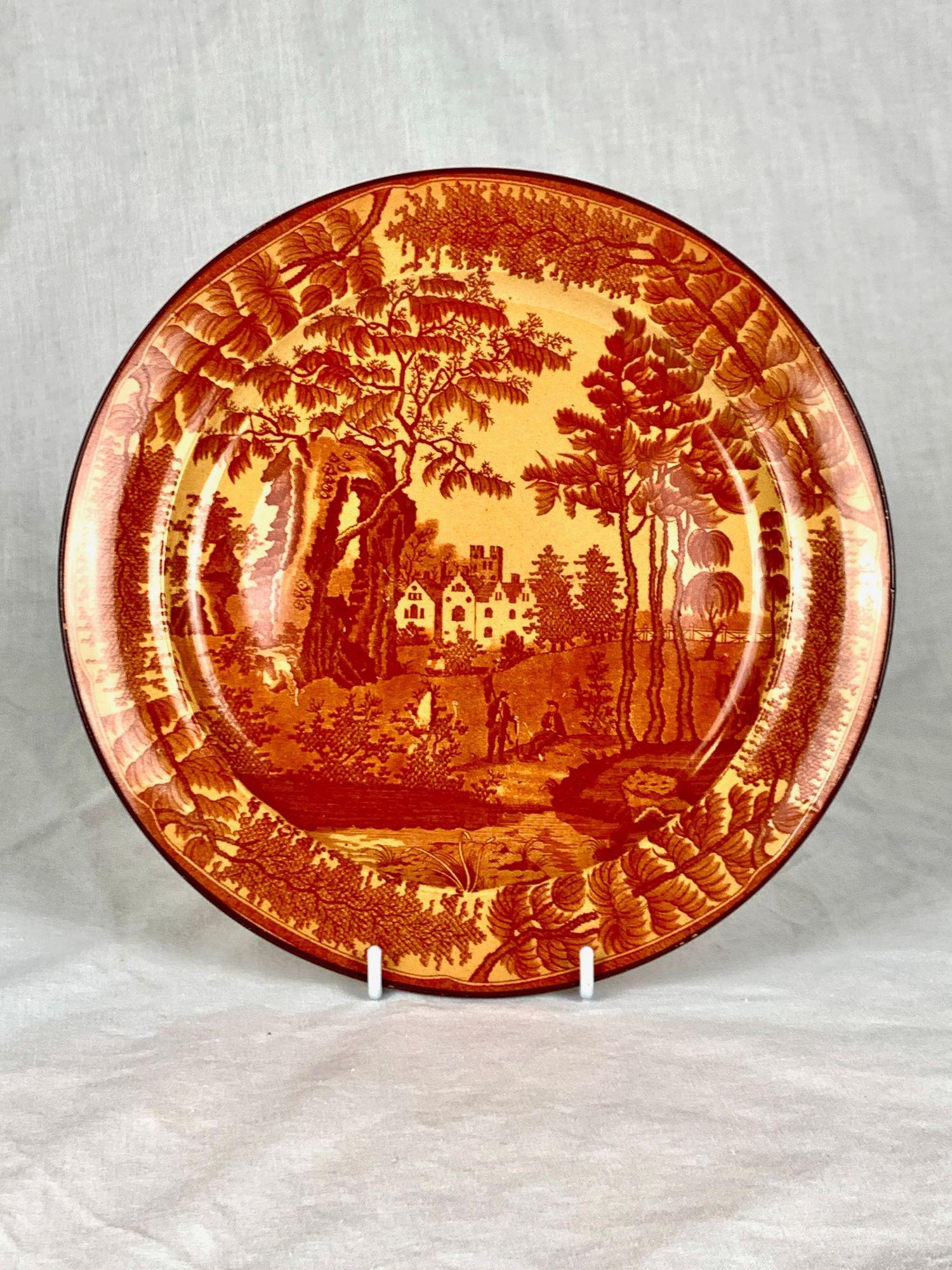This antique English pottery plate, made at Don Pottery circa 1825, is crafted from buff earthenware with a clear glaze that enriches its warm, earthy tone.
The surface is printed in red with a scenic country landscape: tall trees frame a distant
