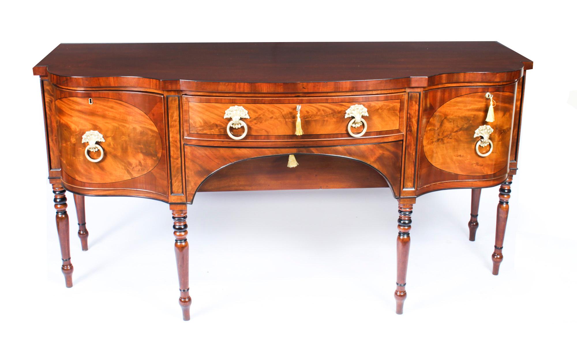 This is a superb antique English Regency flame mahogany shaped serpentine bowfront sideboard, Circa 1820 in date.

It is crossbanded, bordered with ebony lines and fitted with a central frieze drawer above a recessed arched apron drawer. It is