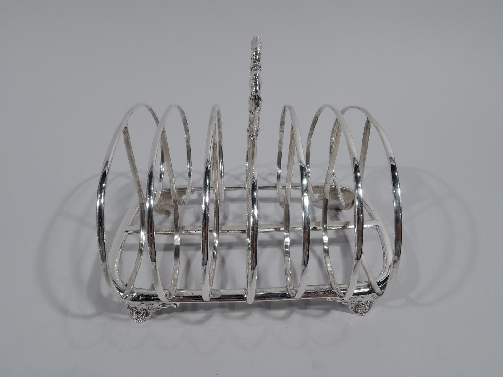 William IV sterling silver toast rack, 1835. Seven double-hooped stretchers mounted to curved and rectangular frame with central stretcher. Reeded ring finial with tooled leaves and lion’s heads. Four leaf-mounted scroll supports. Fully marked