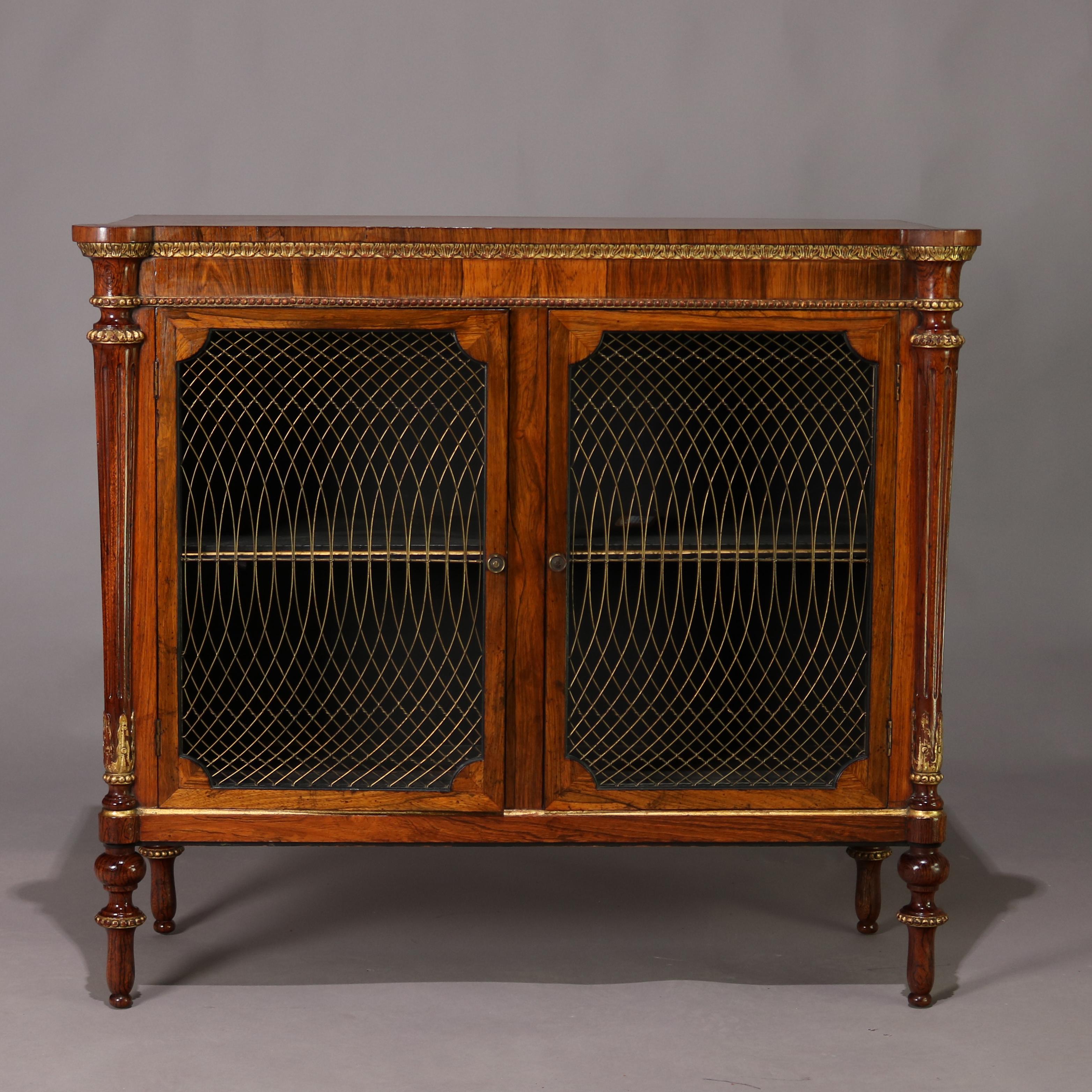 An antique English Regency credenza features rosewood construction with shaped top surmounting cabinet with foliate gilt trim and double bronze mesh doors opening to shelved interior and flanked by gilt decorated columns raised on turned and tapered