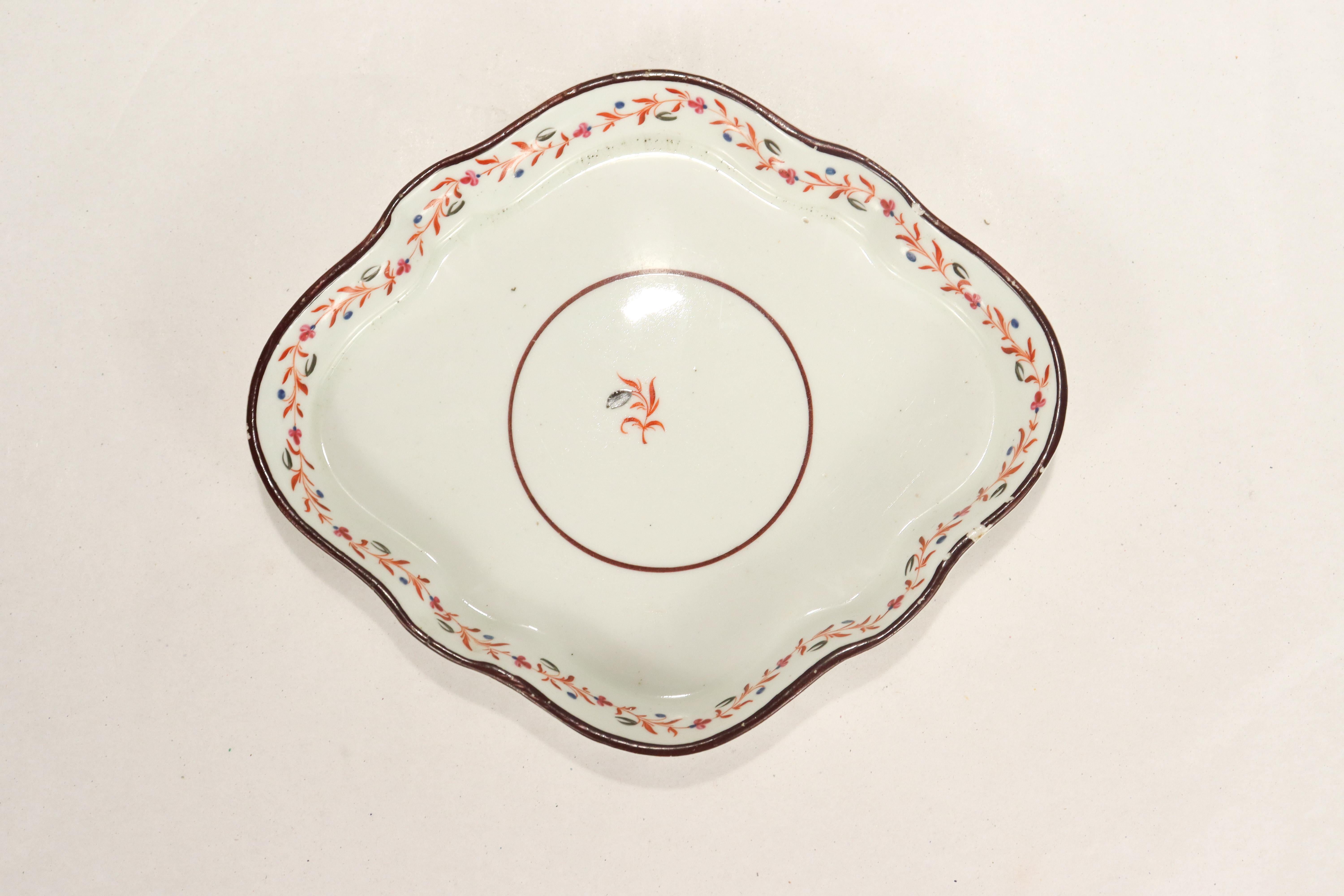 A fine antique English porcelain teapot trivet. 

Attributed to New Hall.

In a diamond-shaped form.

With a single red & black floral sprig inside of a circle to the center. The rim is decorated with a floral garland and a brown painted edge.
