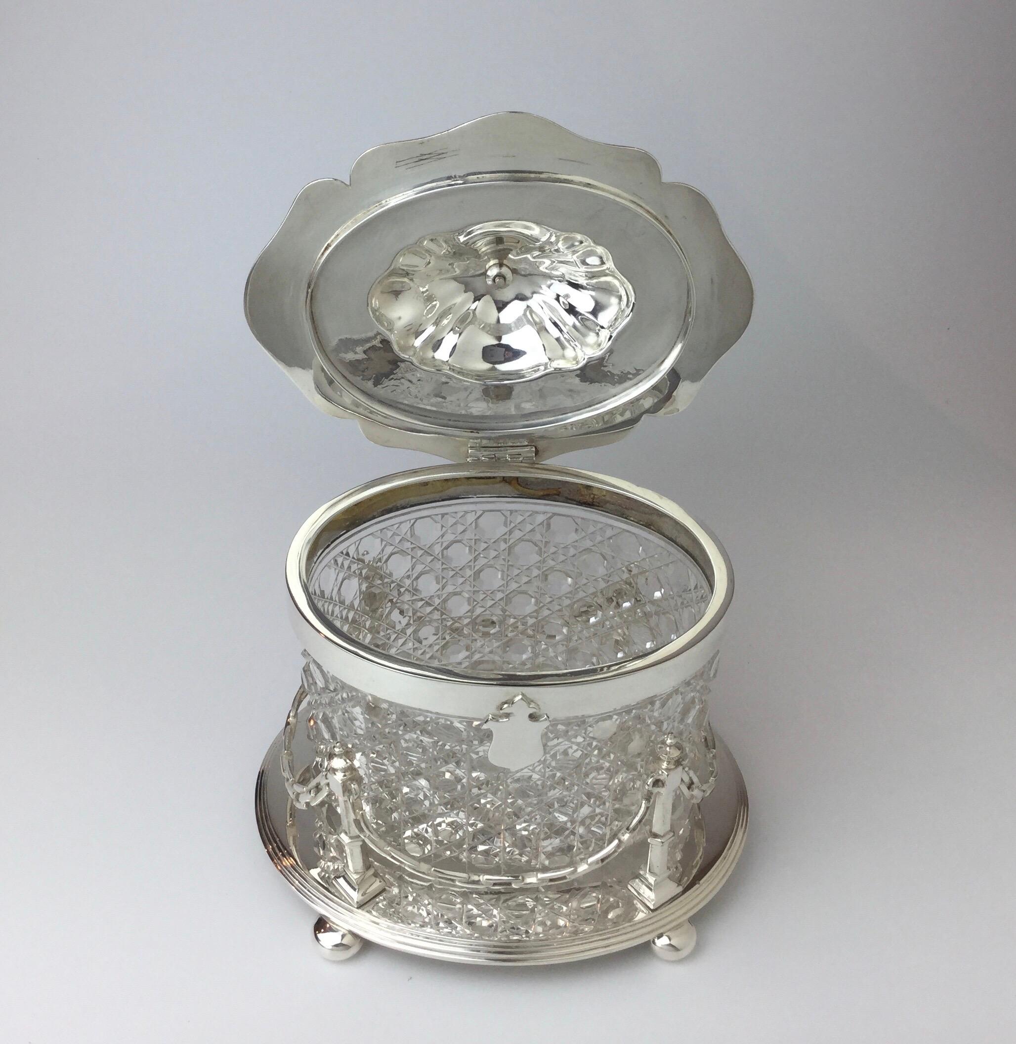 Elegant English biscuit box with cut glass insert. The lid with a tapered finial handle and the body with elongated chain and post design. The oval stepped base resting on four bun feet.