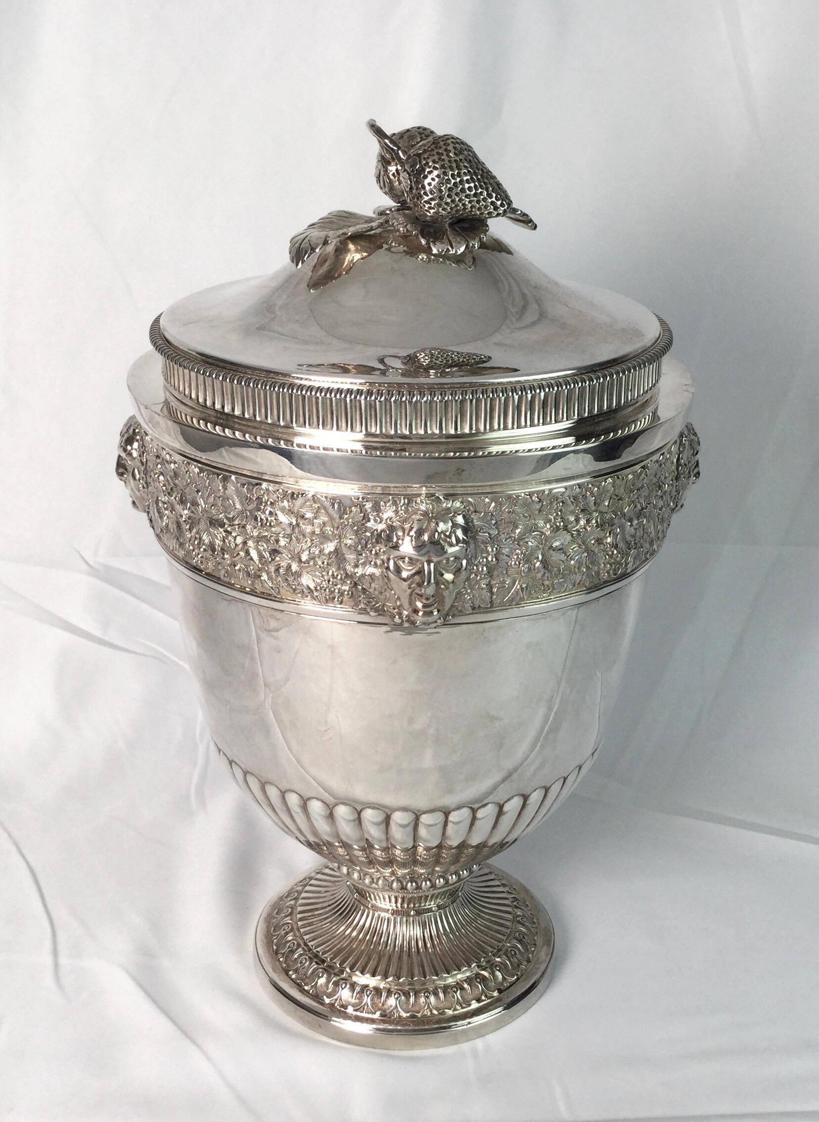 A classic urn form ice bucket or wine cooler with a liner, England, circa 1900. The urn form with a finial on the lid of strawberries and leaves, the body at the top with cast decoration of grape leaves and masks of bacchanalia. The pedestal base