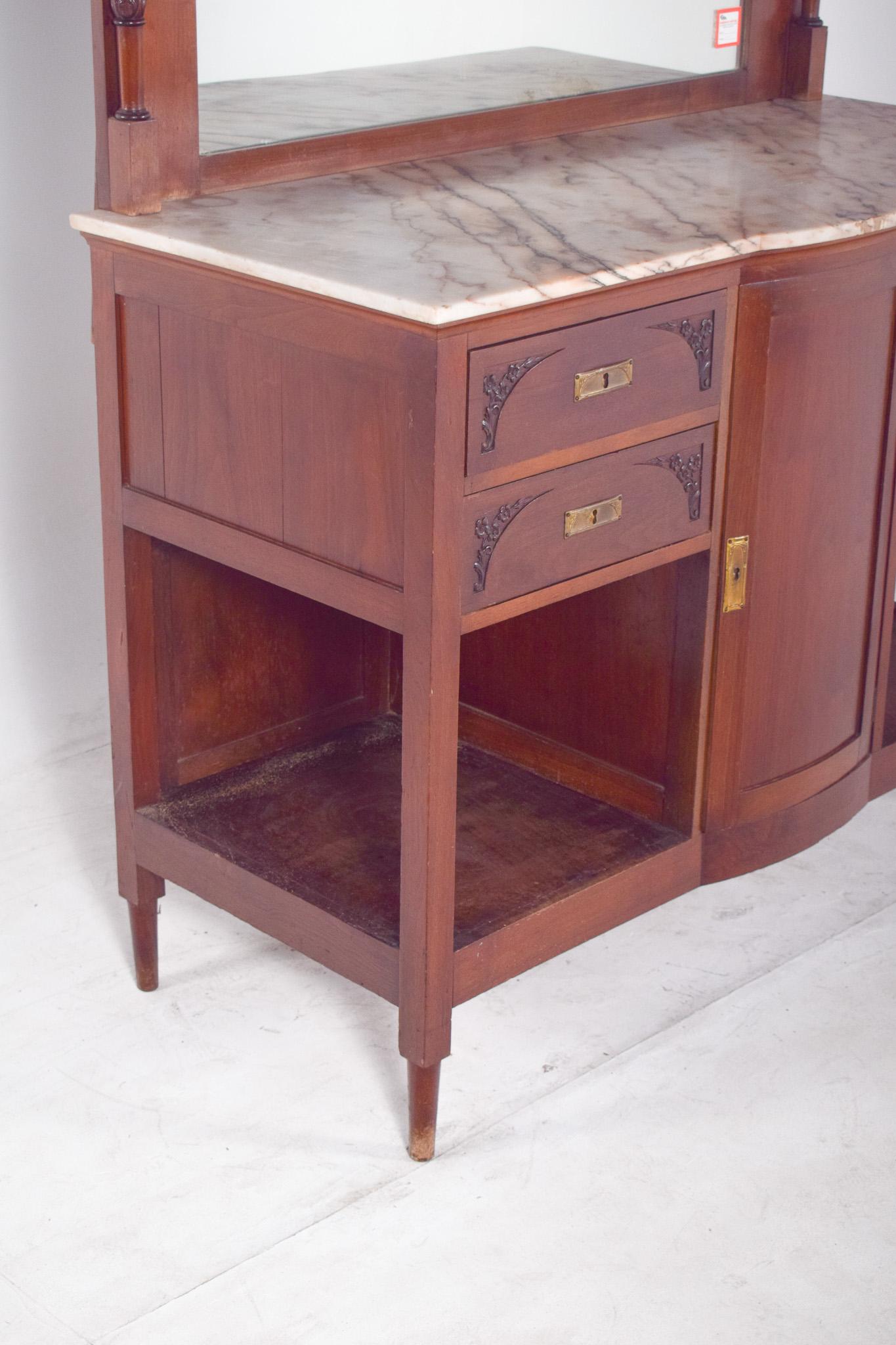 Sideboard with mirror and marble top. The upper structure houses a rectangular mirror flanked by small turned columns, crowned by a central decorative carving in the shape of a bow, which accentuates the ornamental character of the piece.
The lower