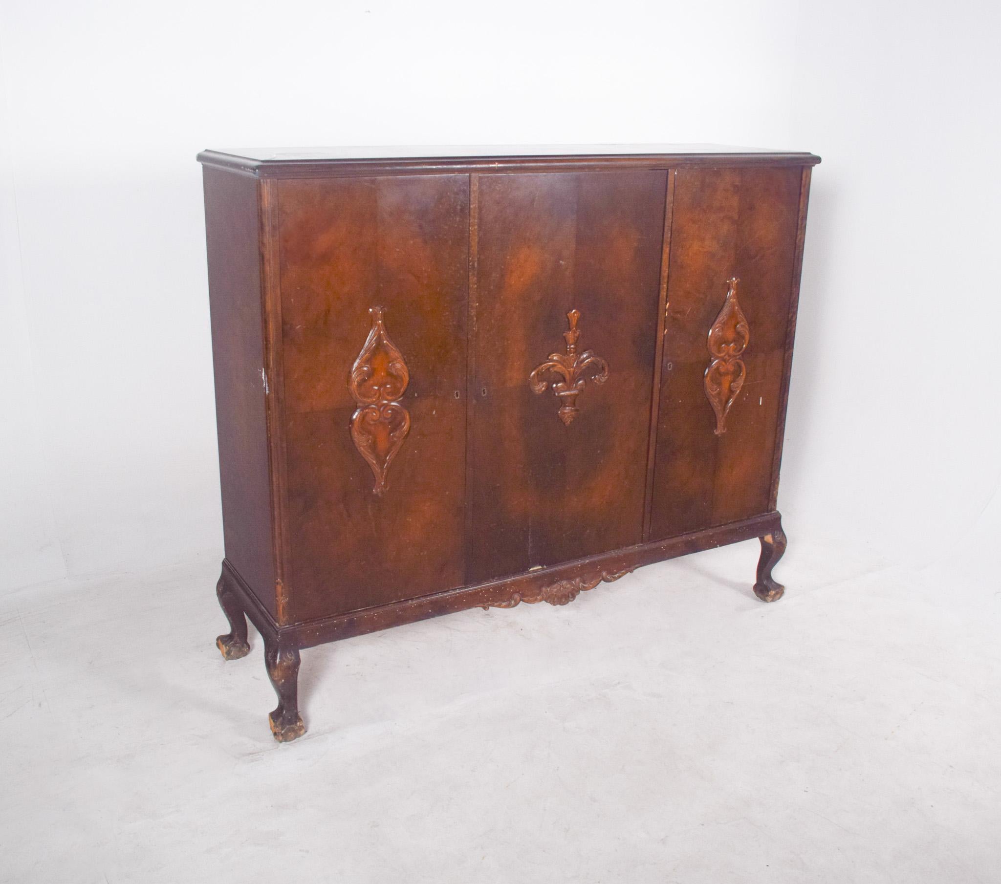 Credenza a tre ante. In legno scuro, con ripiani all'interno, ha una struttura robusta e simmetrica, con tre porte anteriori che presentano intagli centrali in rilievo, di cui quella centrale è particolarmente elaborata, con un motivo che ricorda un