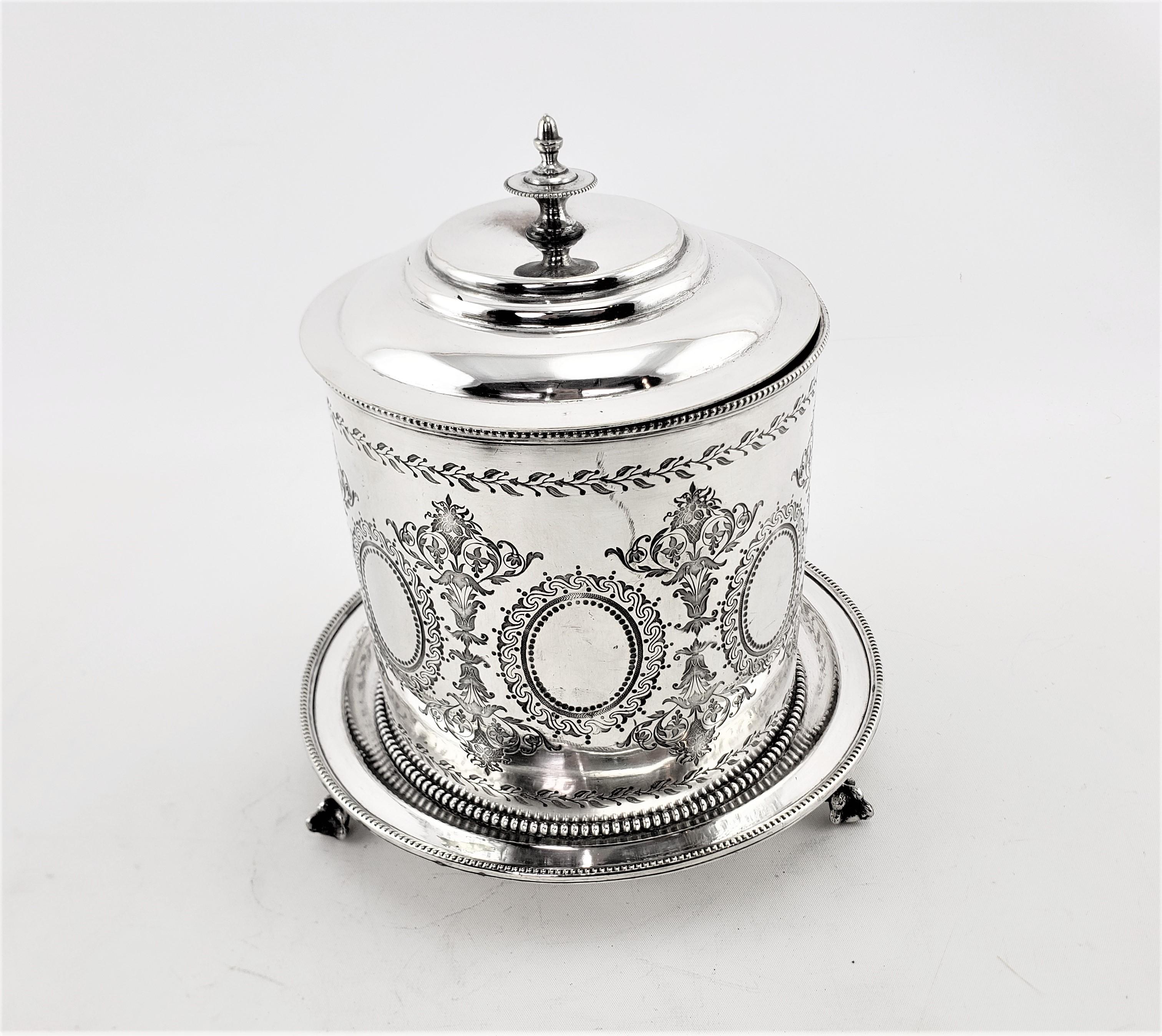 This antique oval shaped and silver plated biscuit barrel was made in England in approximately 1880 in a period Victorian style. This biscuit barrel has an oval shape with a hinged lid and elaborate stylized wreath engraving around the sides and