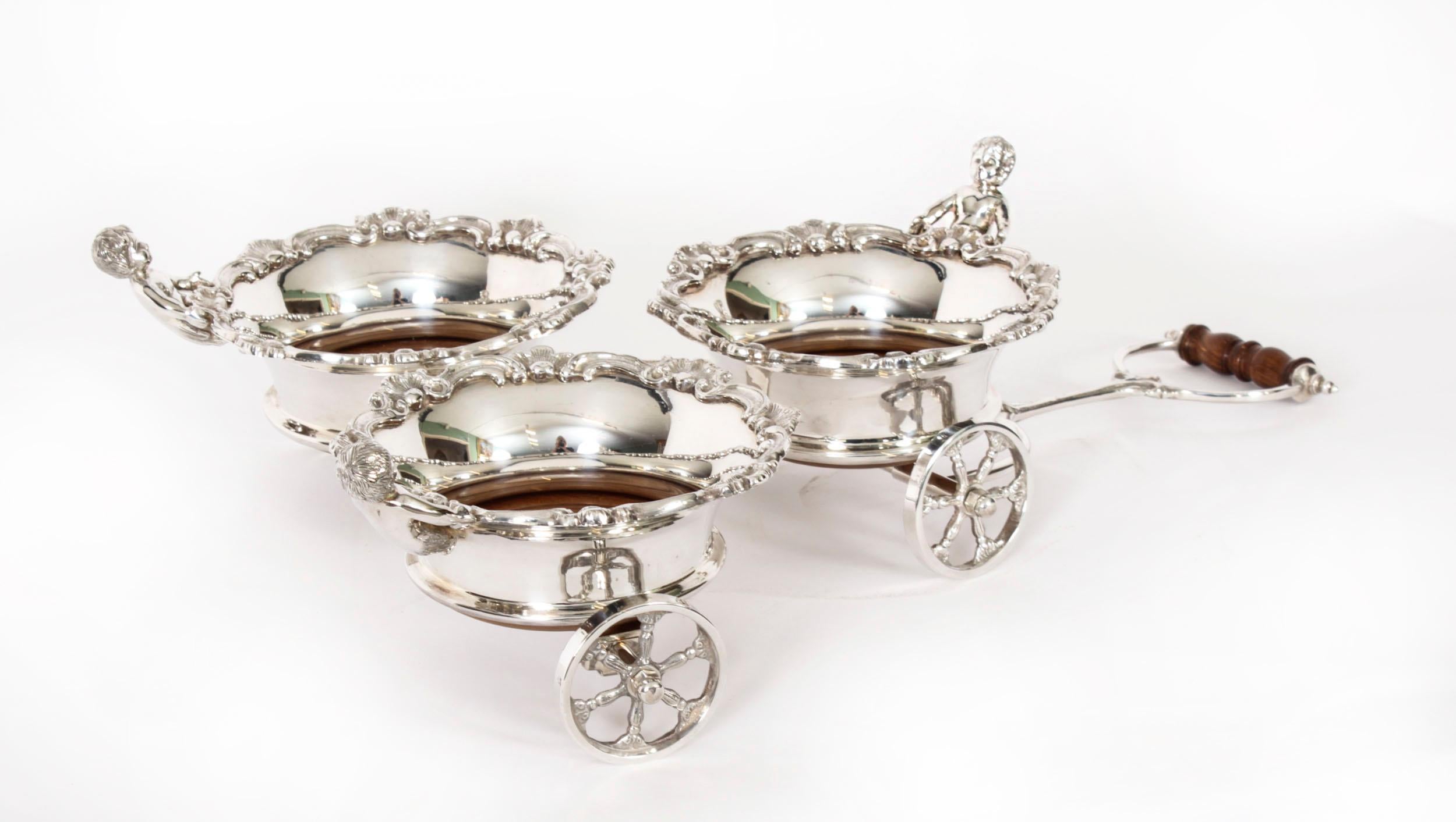 This is a superb antique English silver plated triple bottle coaster, circa 1930 in date.
 
The three coasters are arranged in a triangle on wheels, each are decorated with an acanthus rim and a cherub, the handle at the front steers the coaters.
