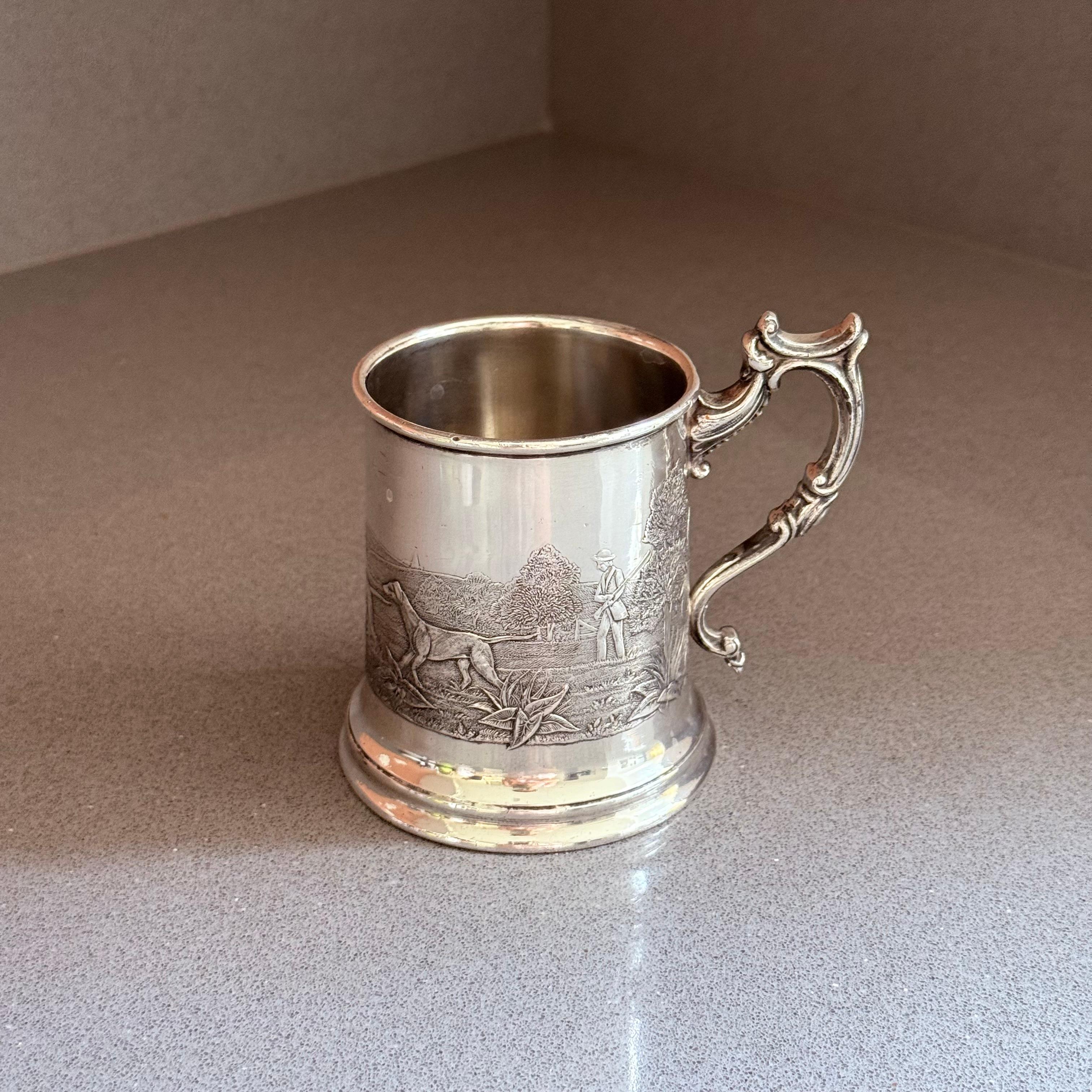 Cattura l'eleganza dell'artigianato tradizionale inglese con questo boccale antico placcato in argento, splendidamente decorato con una scena di caccia incisa con uno sportivo, un cane e un paesaggio naturale. Il manico ornato e i rilievi finemente