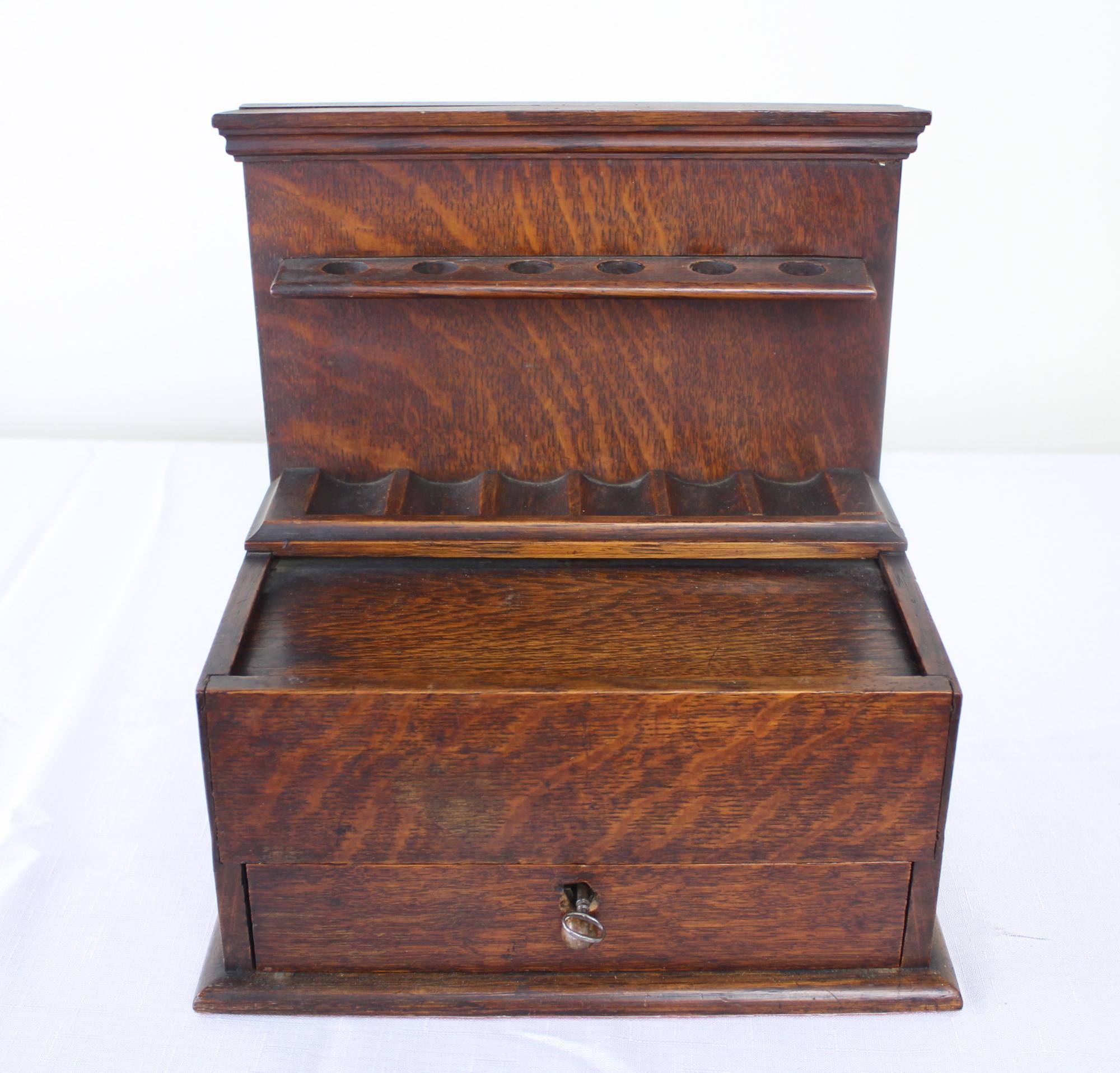 A finely detailed English oak smoking box. At the top are six charming holes for pipes, while the drawer below, with working key, opens to reveal several compartments for cigarettes and accessories. The pull action of the drawer simultaneously opens