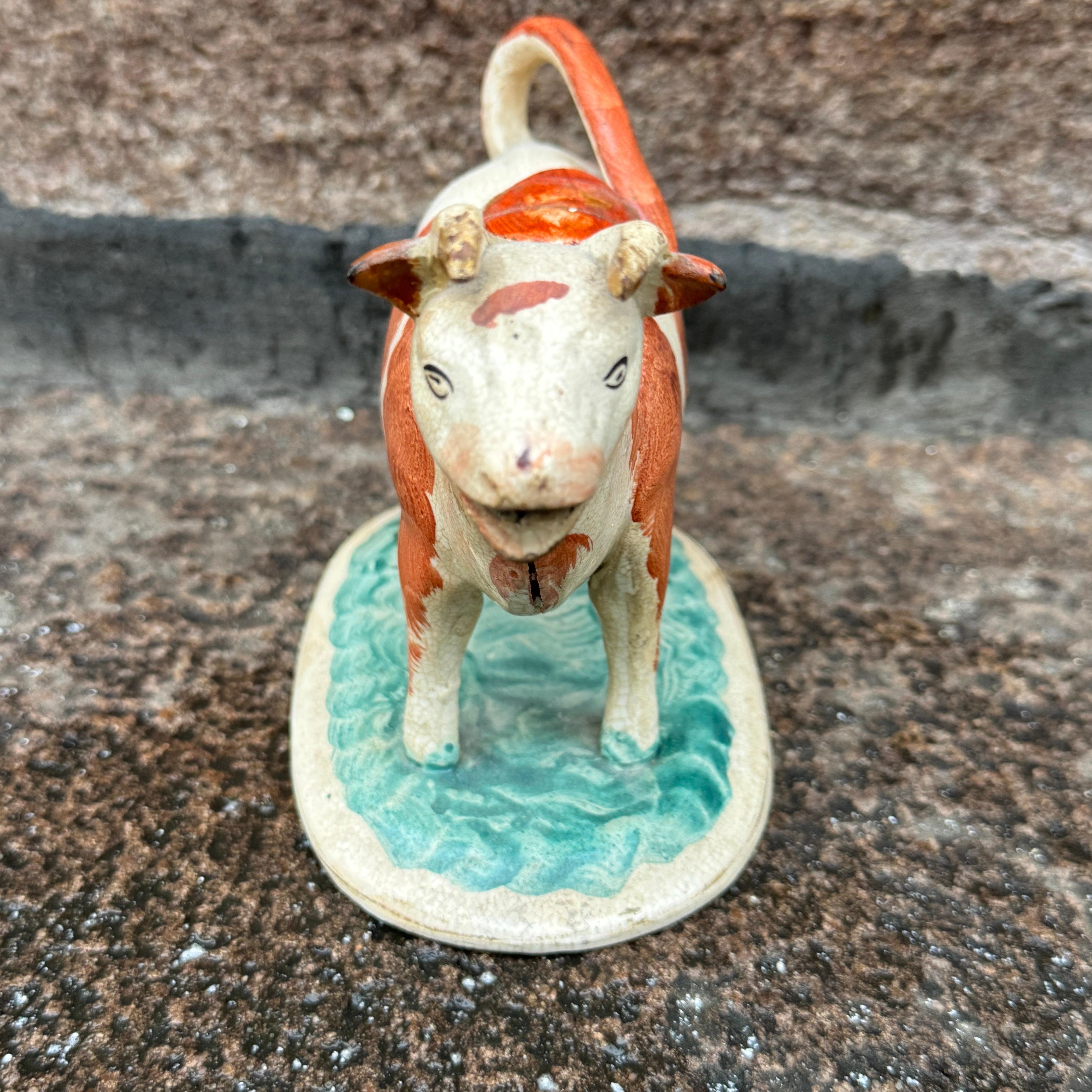 Encantadora cremera de vaca o toro de cerámica inglesa antigua de Staffordshire de mediados del siglo XIX. La dócil criatura, cuya cola forma el asa, se yergue pacientemente sobre una base cubierta de hierba. Esta pieza conserva su botón original.