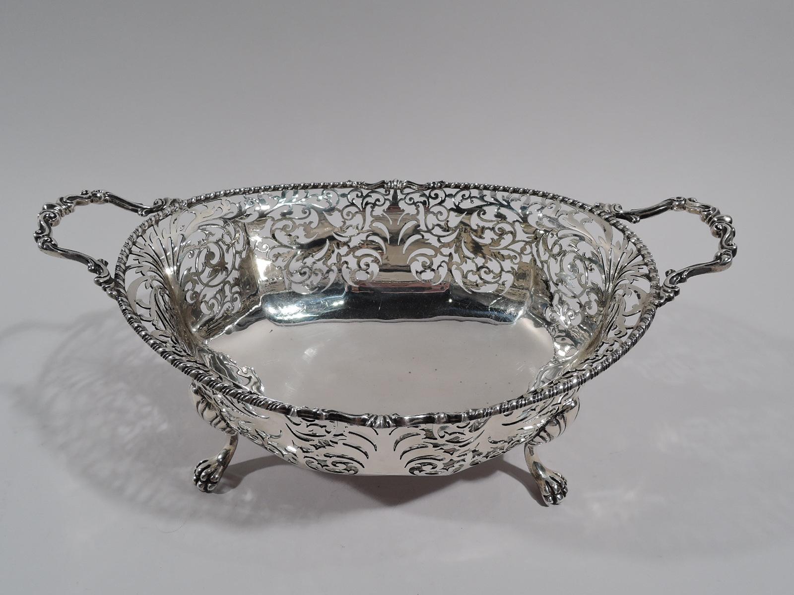 George V sterling silver basket bowl. Made by Mappin & Webb in London in 1919. Oval with solid and faceted well. Sides have pierced ornament: Fluid and symmetrical scrolls and foliage. Cabled rim with scrolled bracket handles. Four paw supports with