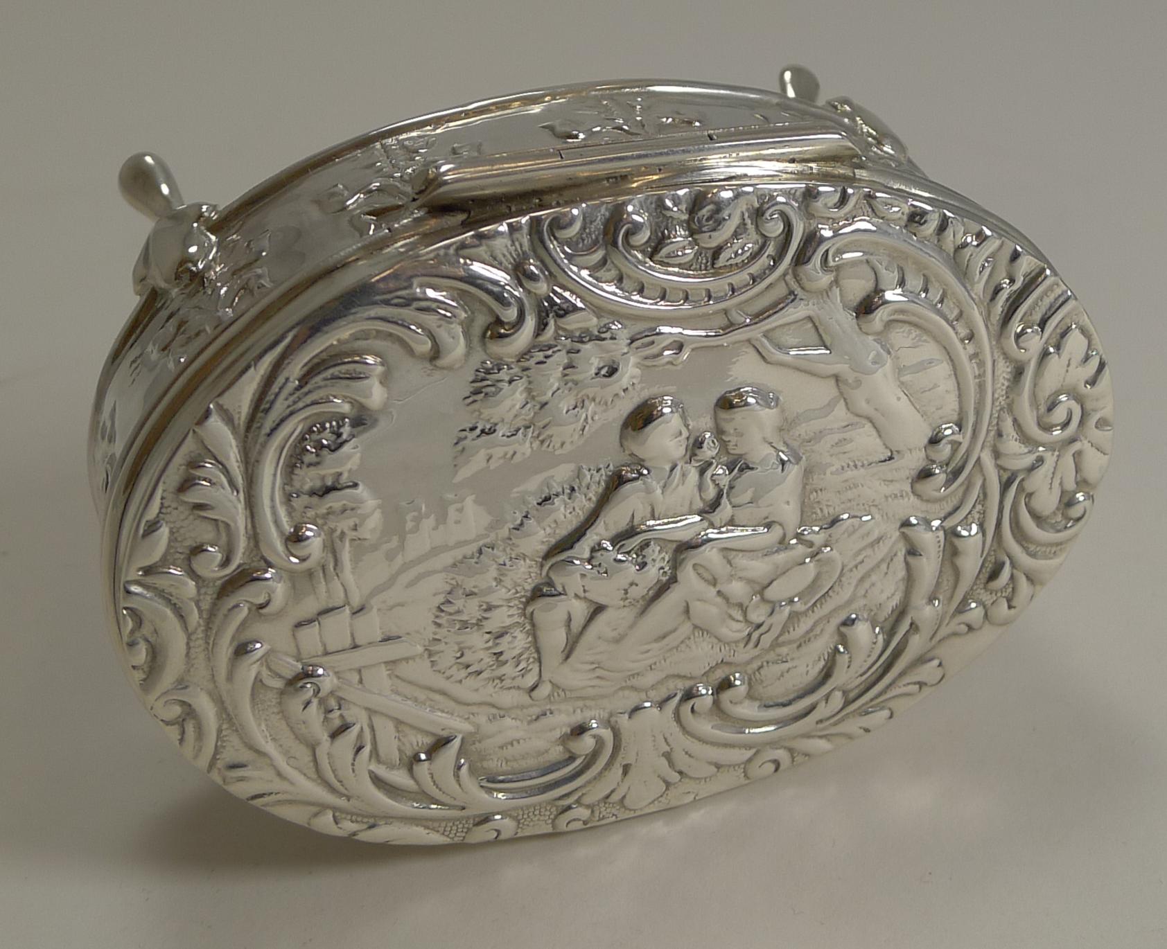 Pretty as a picture, this Edwardian oval sterling silver box stands on four elegant legs. The sides have an embossed or repousse decoration featuring a series of ribbon and bow motifs.

The lid is beautifully decorated with a romantic courting