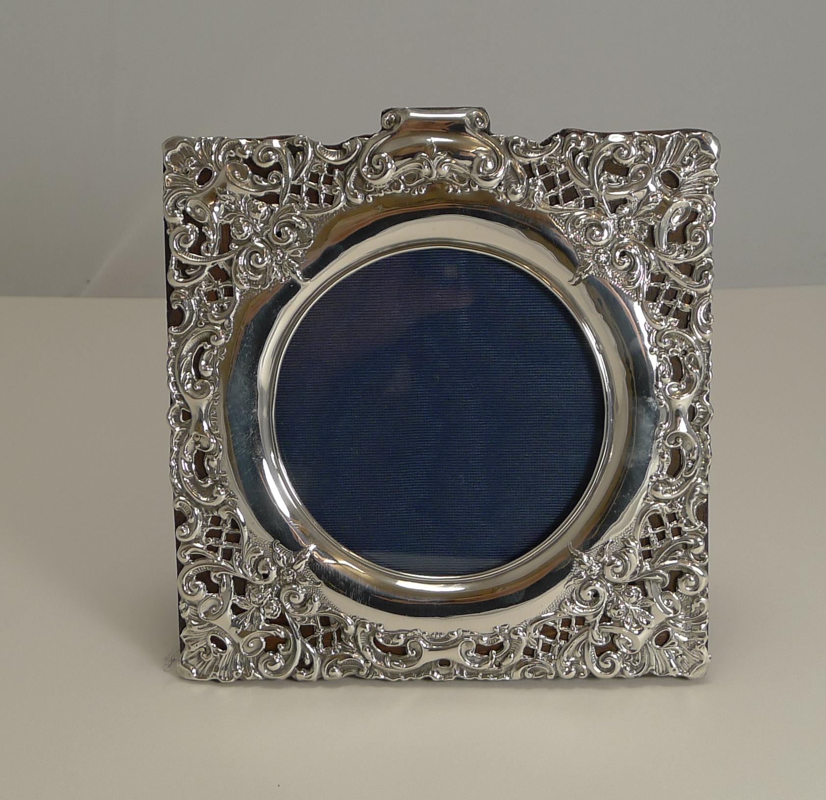 A stunning antique English sterling silver frame of square form with a circular glazed aperture.

The back is made from solid English oak and incorporates a folding easel stand. The intricate and highly decorative silver is topped with a scroll