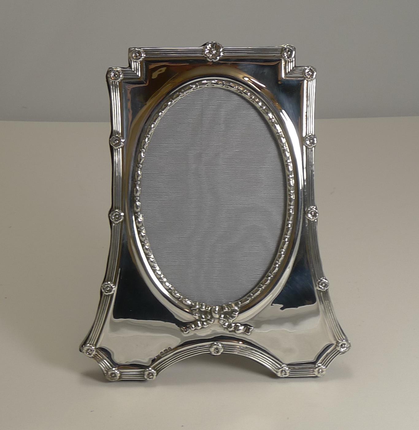 A beautifully shaped photograph frame made from English sterling silver with a solid oak backing with a folding easel stand.

The edge is decorated with a linear design punctuated with floral motifs; the oval aperture is surrounded with a raised