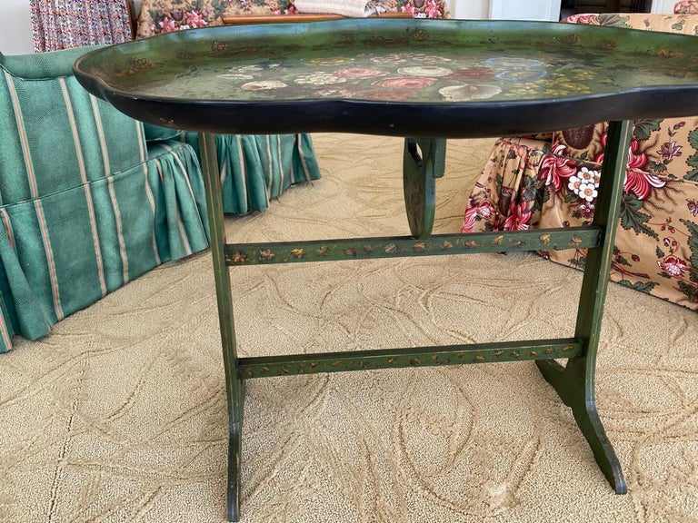 The antique tole antique tray coffee table features a scalloped tray hand painted of flowers on a green background with gold leaf and having a green wooden stand to match, Early 19th century.